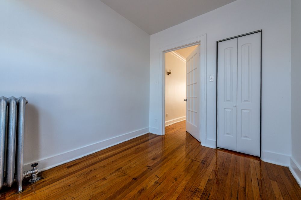Empty room with hardwood floors, white walls, closet, doorway to hallway, and radiator.