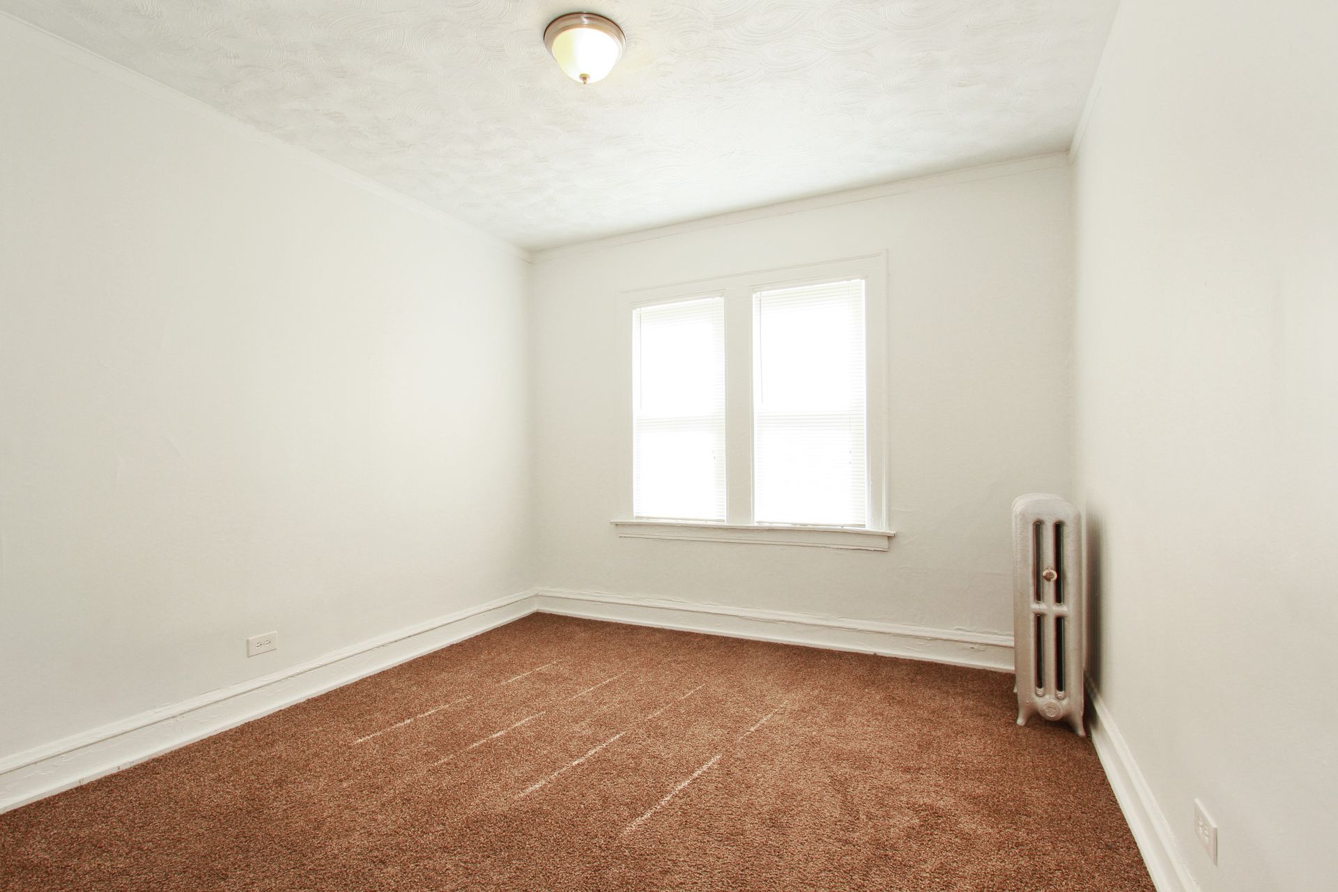 Empty room with brown carpet, window, white walls, radiator, and ceiling light.