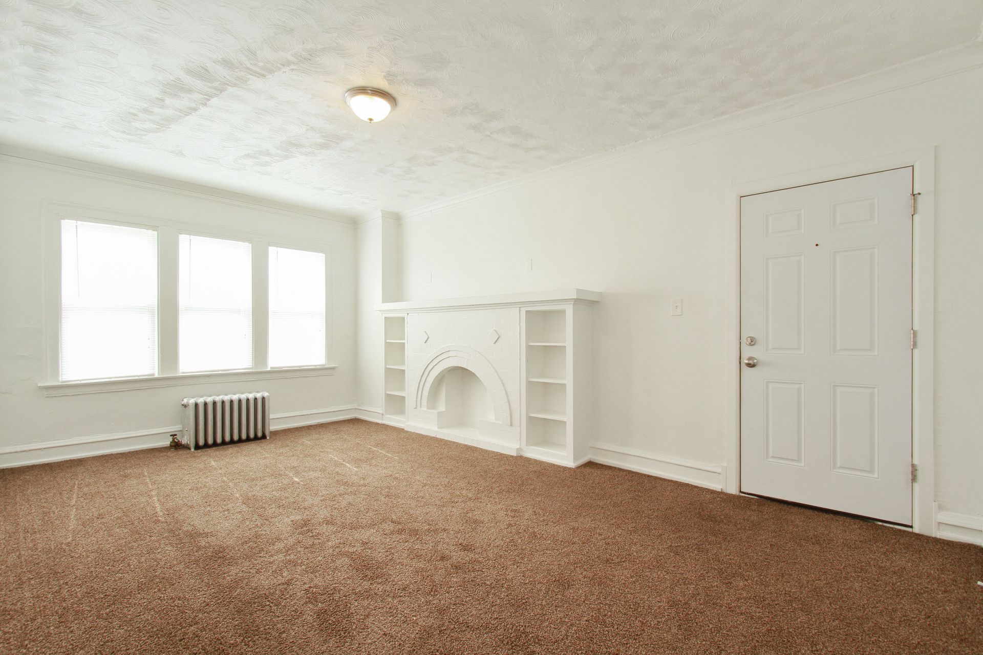 Empty living room with brown carpet, white walls, built-in fireplace, and large windows.