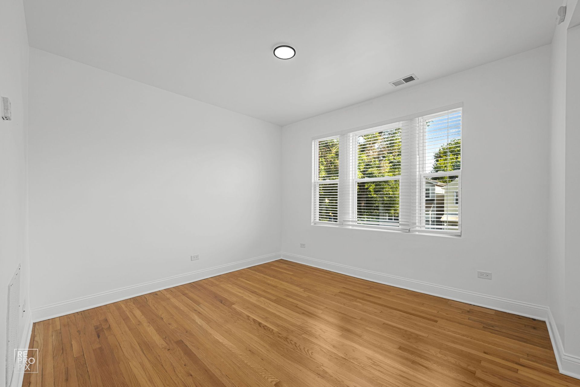 Empty room with hardwood floors, white walls, and a triple window with blinds.