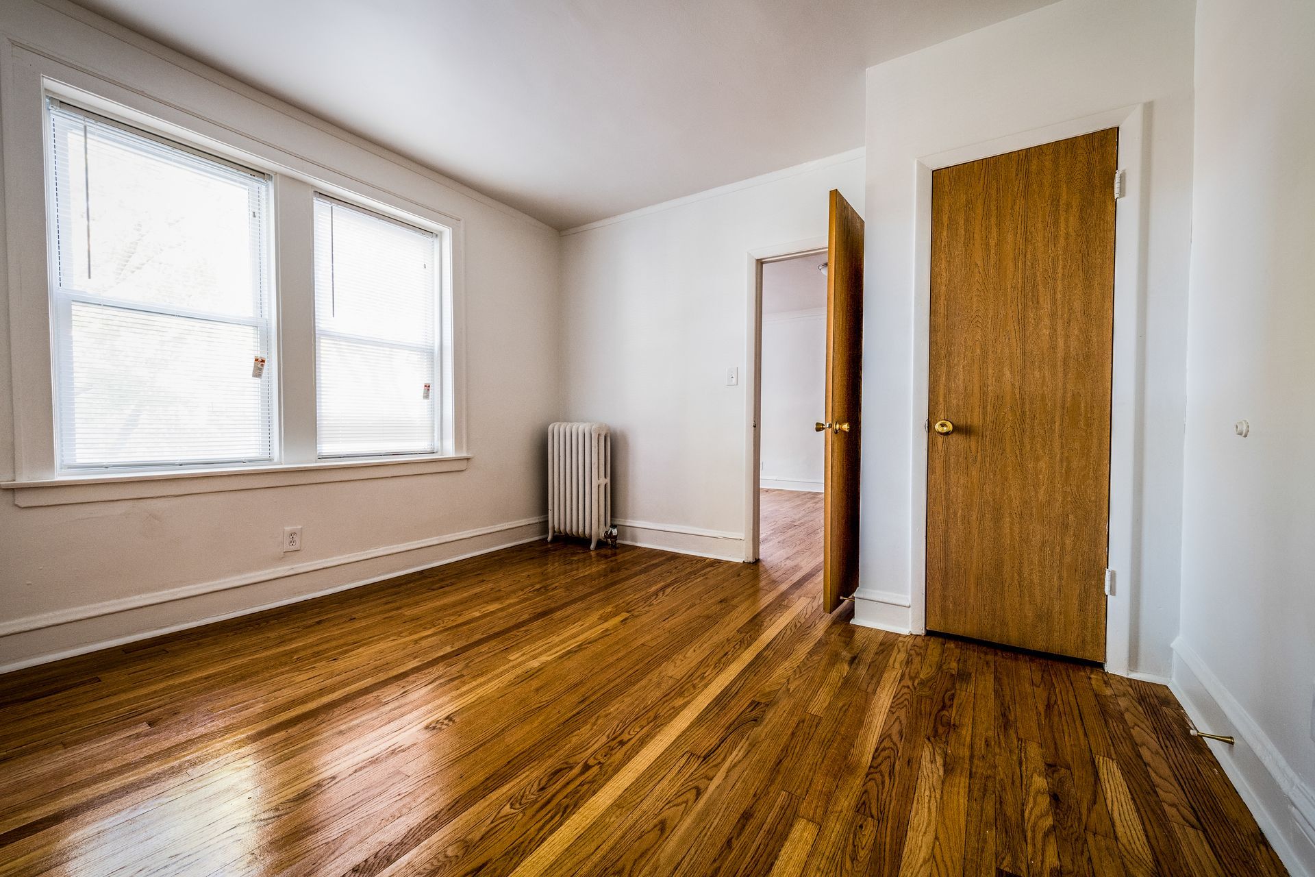 Empty room with hardwood floors, a closed wooden door, and a partially open doorway. White walls and window.