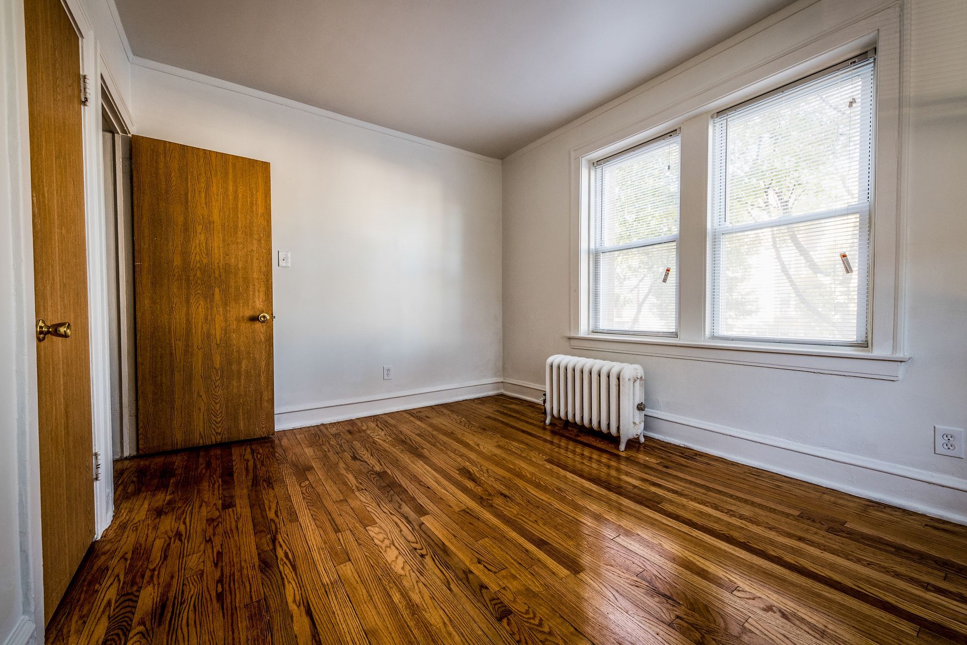 Empty room with wood flooring, door, two windows, and radiator.