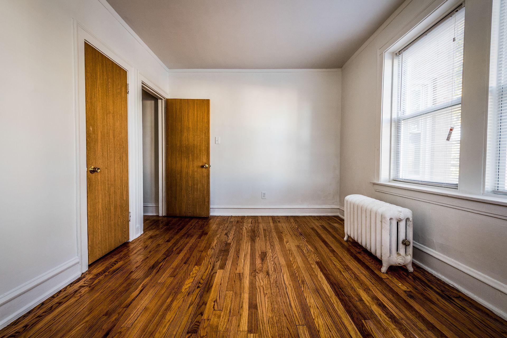 Empty room with hardwood floors, white walls, two wooden doors, and a window with a radiator.