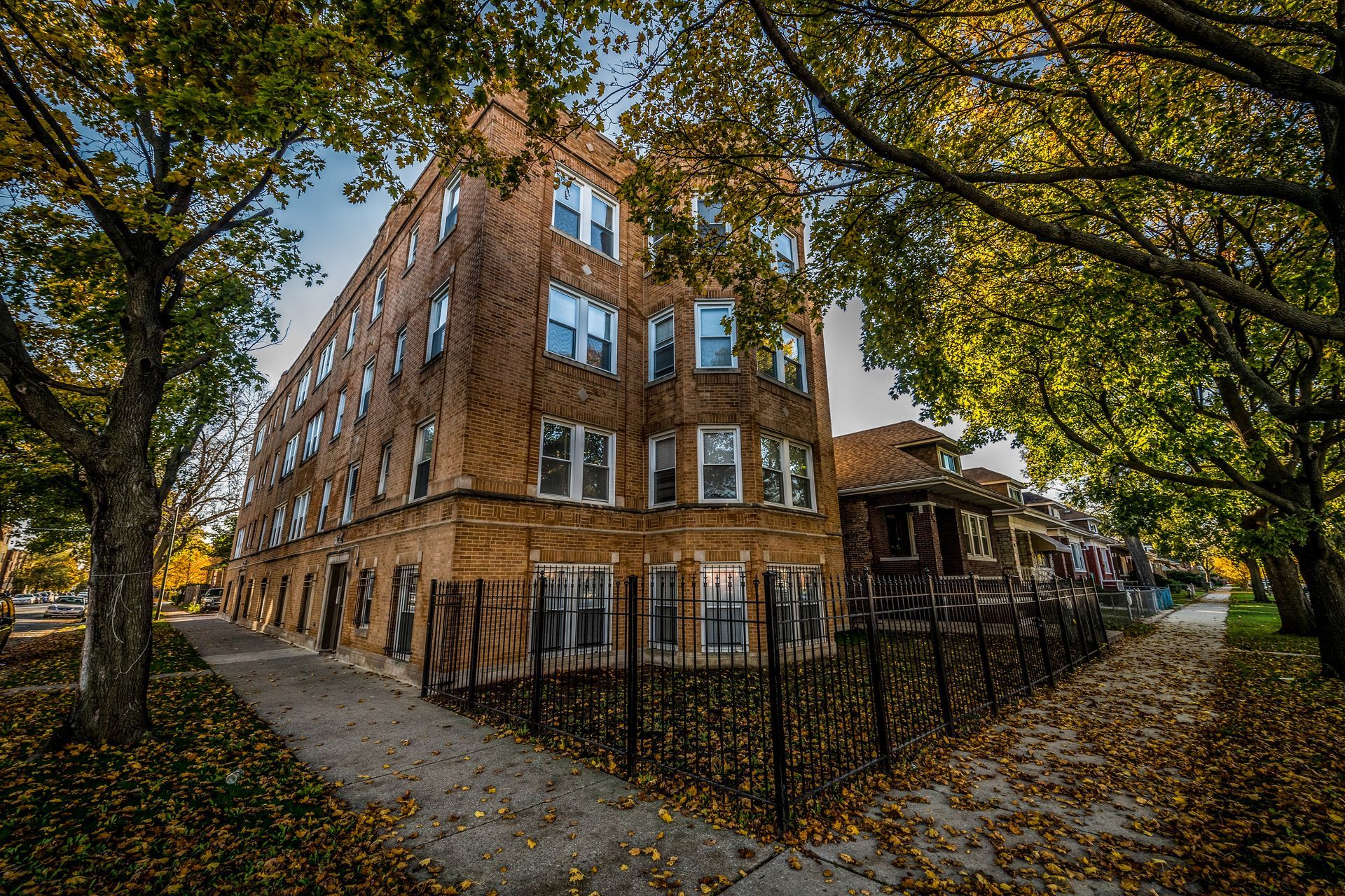 Brick apartment building framed by fall trees on a sidewalk, fence in front.