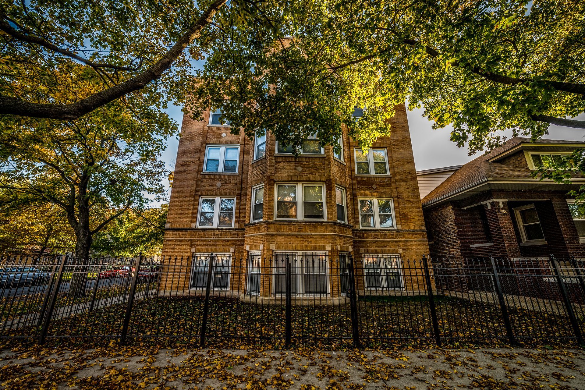 Multi-story brick building with iron fence, autumn leaves, and trees.