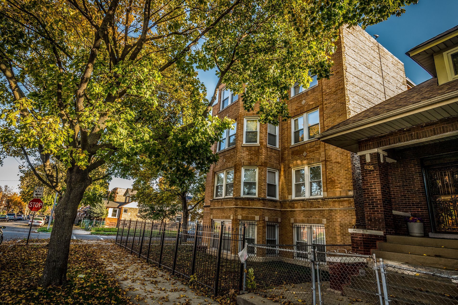 Brick apartment building next to a house, with a tree in front. Fall leaves and fence visible.