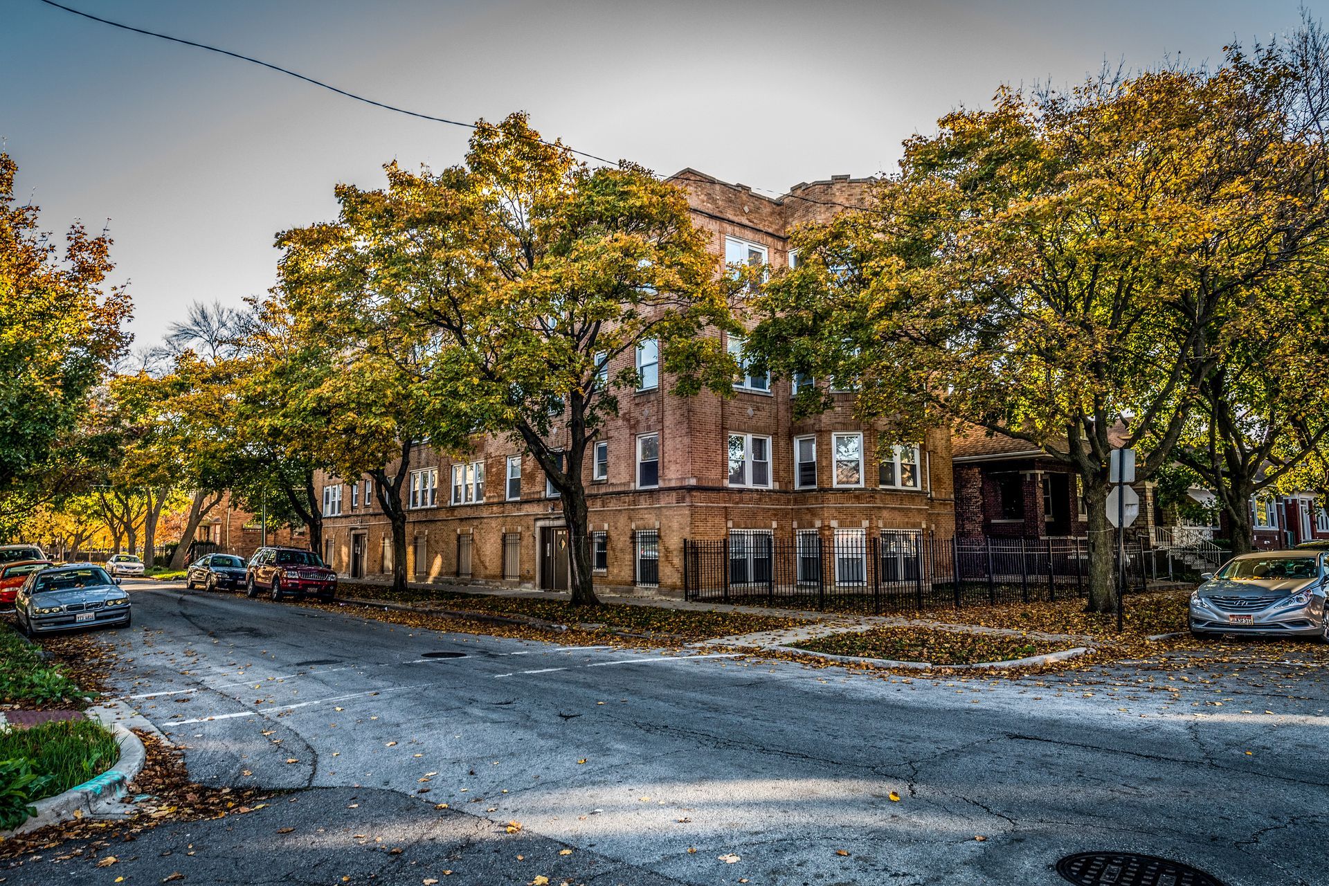 A brick apartment building on a tree-lined street with parked cars and fallen autumn leaves.