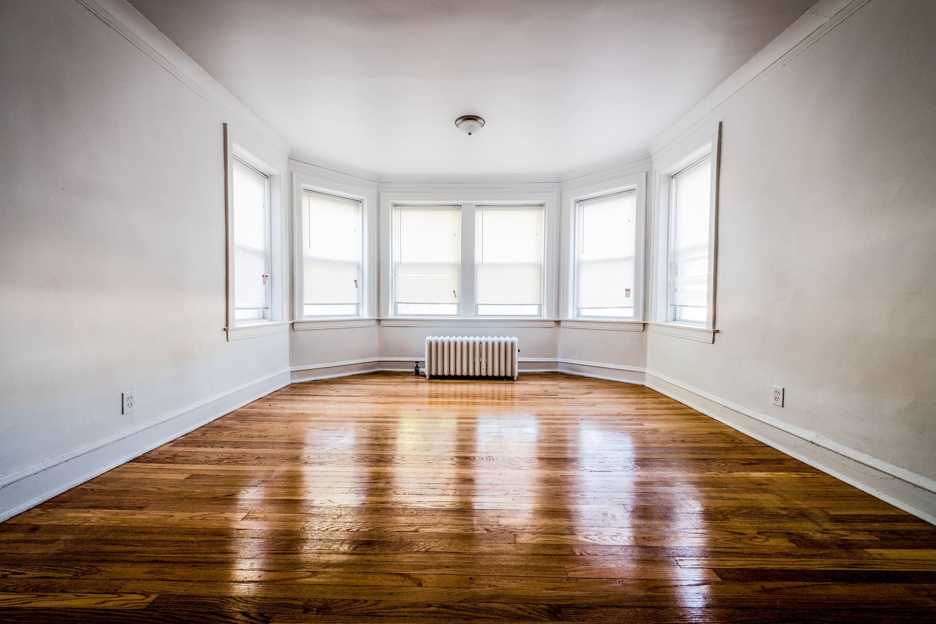 Empty room with hardwood floors, white walls, and a bay window with blinds.