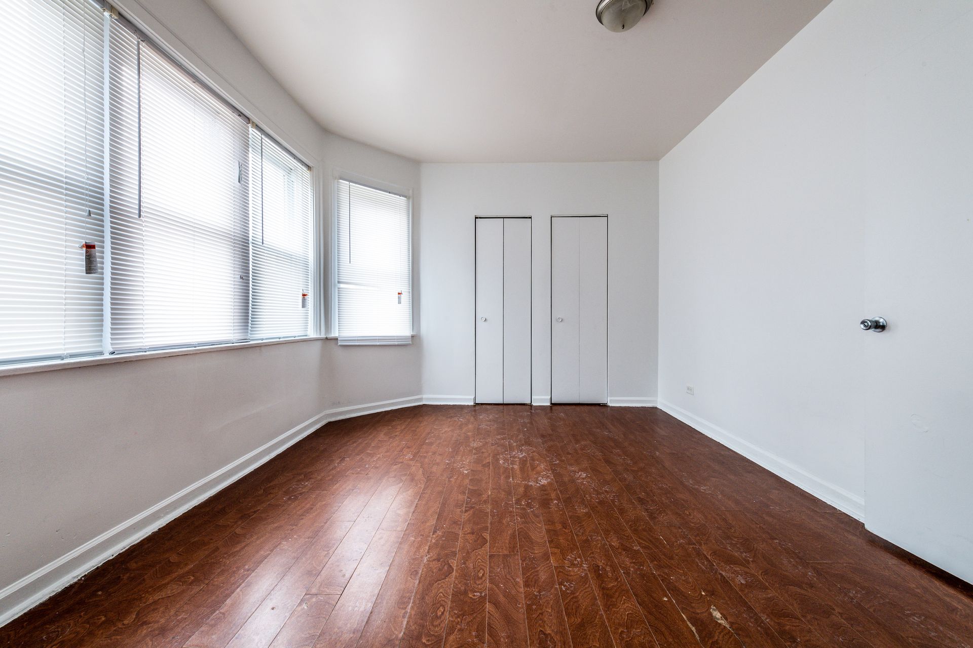 Empty room with hardwood floors, a window with blinds, and a closed door.