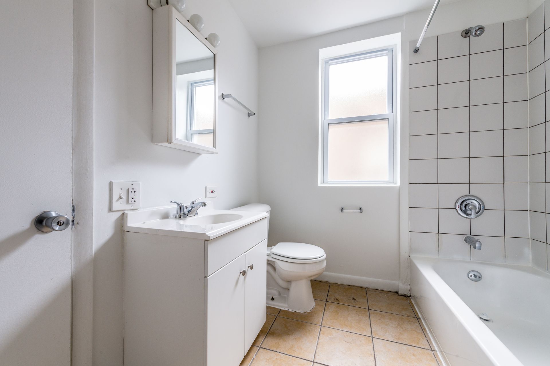 Bathroom with white fixtures: sink, toilet, tub, and window. Beige tile floor.