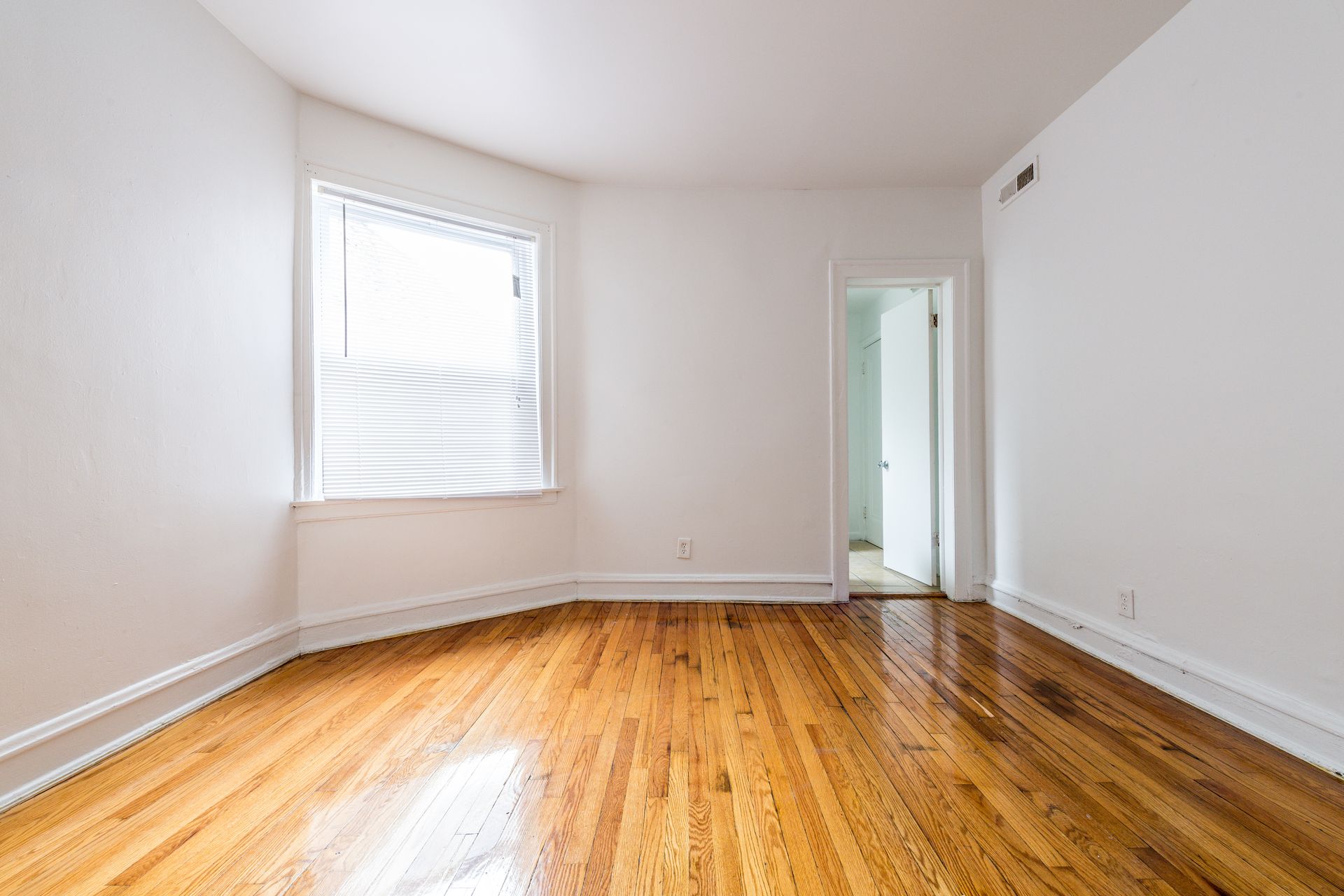 Empty room with hardwood floors, white walls, and a window with blinds. Doorway visible.