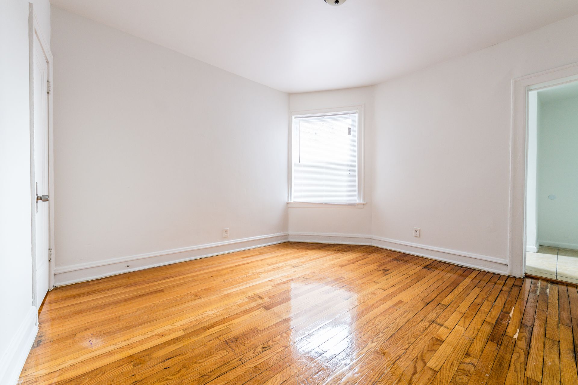 Empty room with hardwood floors, white walls, and a window. A doorway is visible on the right.