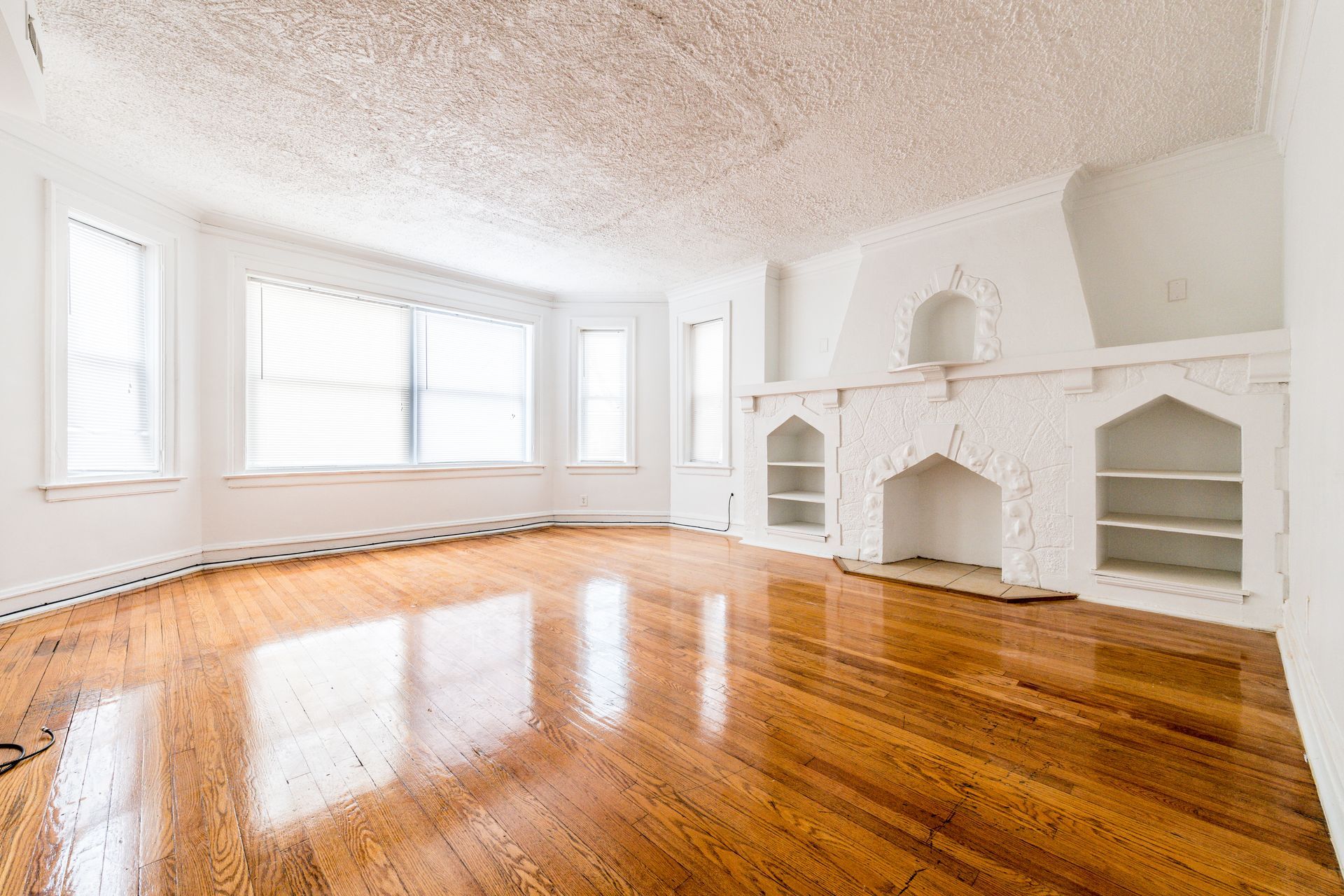 Empty living room with wood floors, white walls, large windows, and a decorative fireplace.