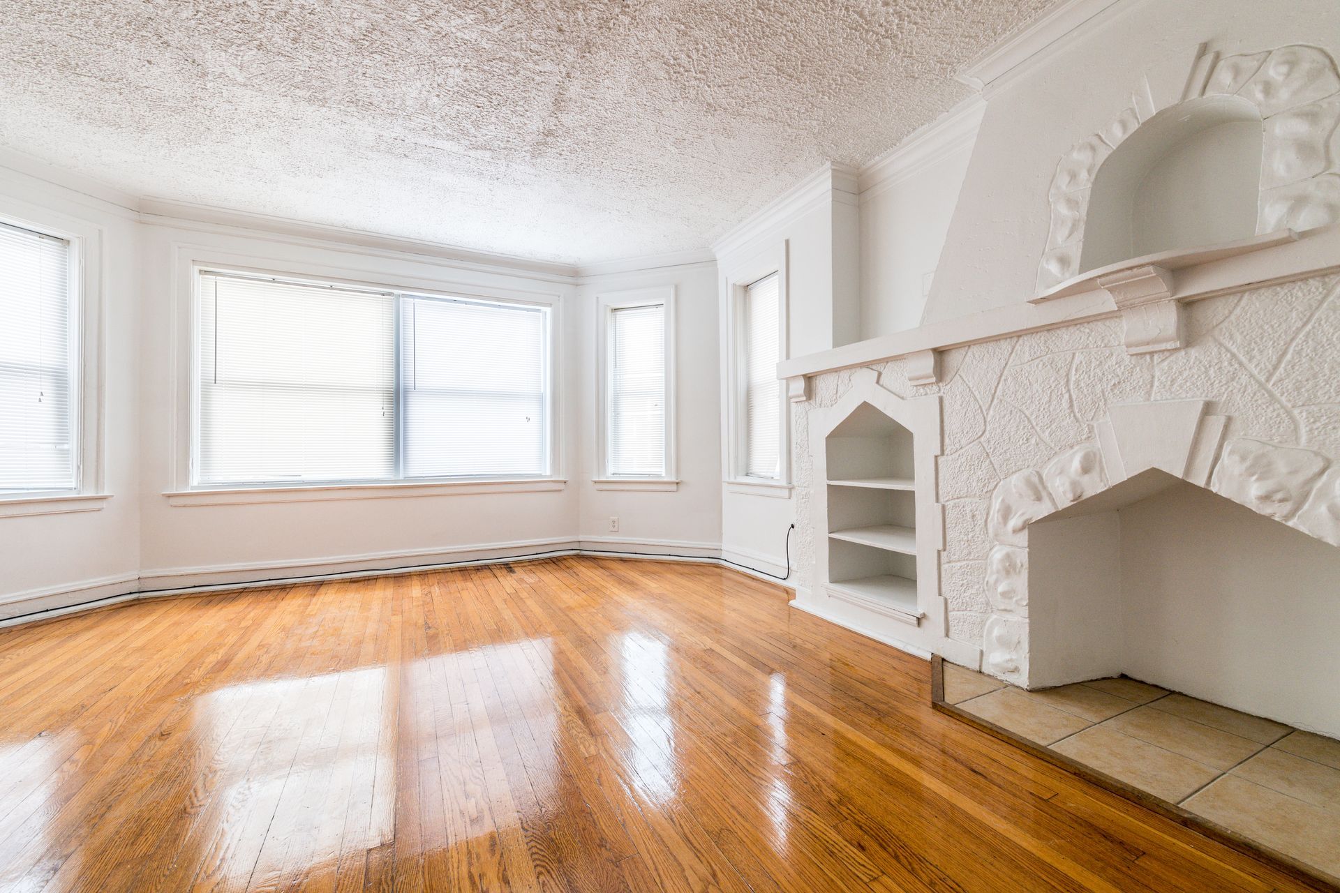Empty, bright living room with wood floors, white walls, ornate fireplace, and large windows.