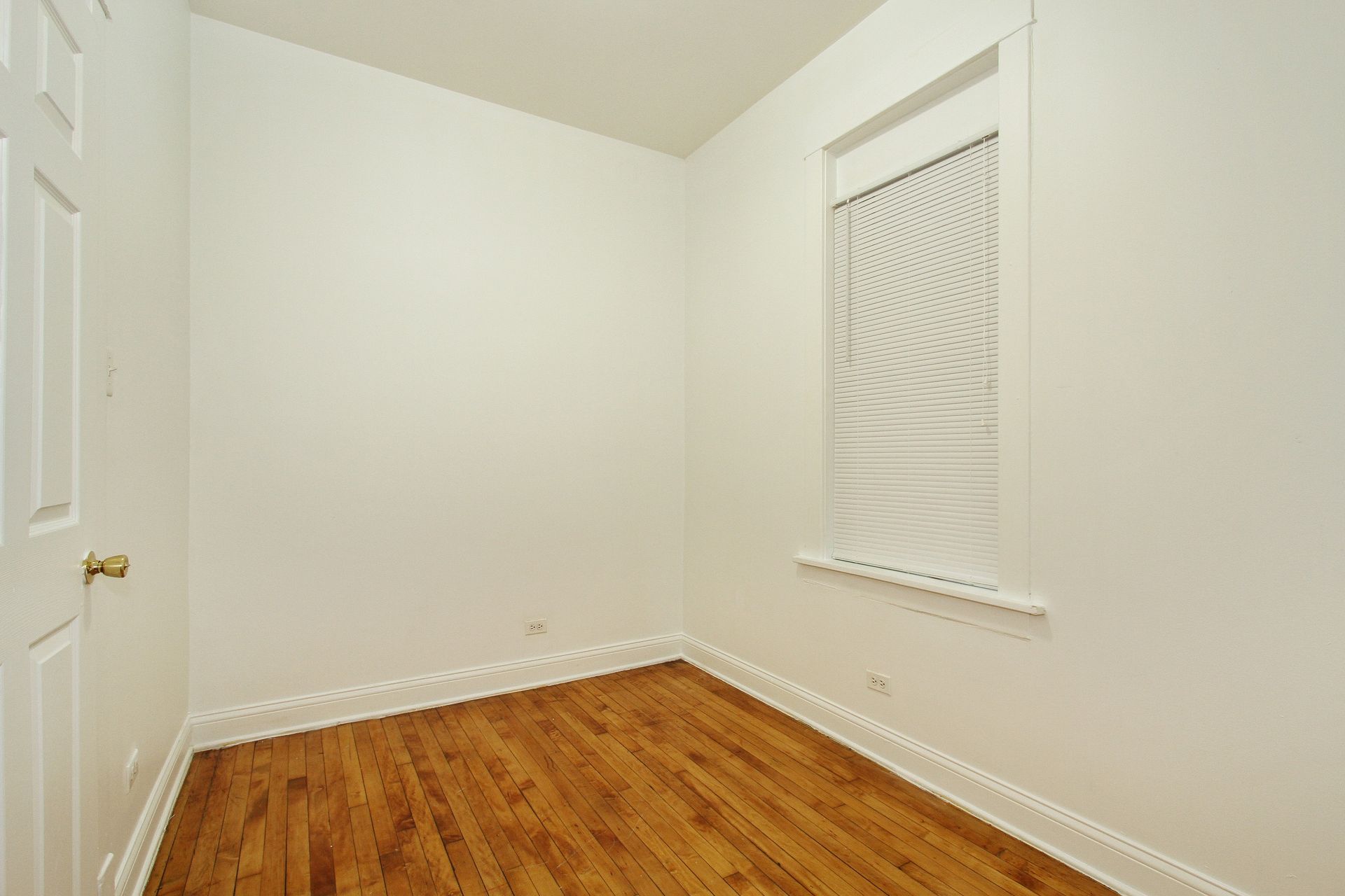 Empty room with hardwood floors, white walls, and a window. A door is on the left.