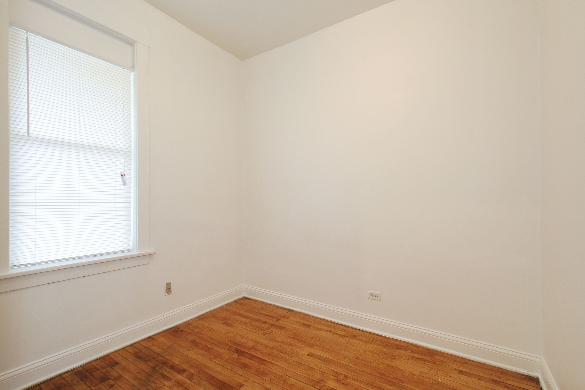Empty room with hardwood floors, a window with blinds, and white walls.