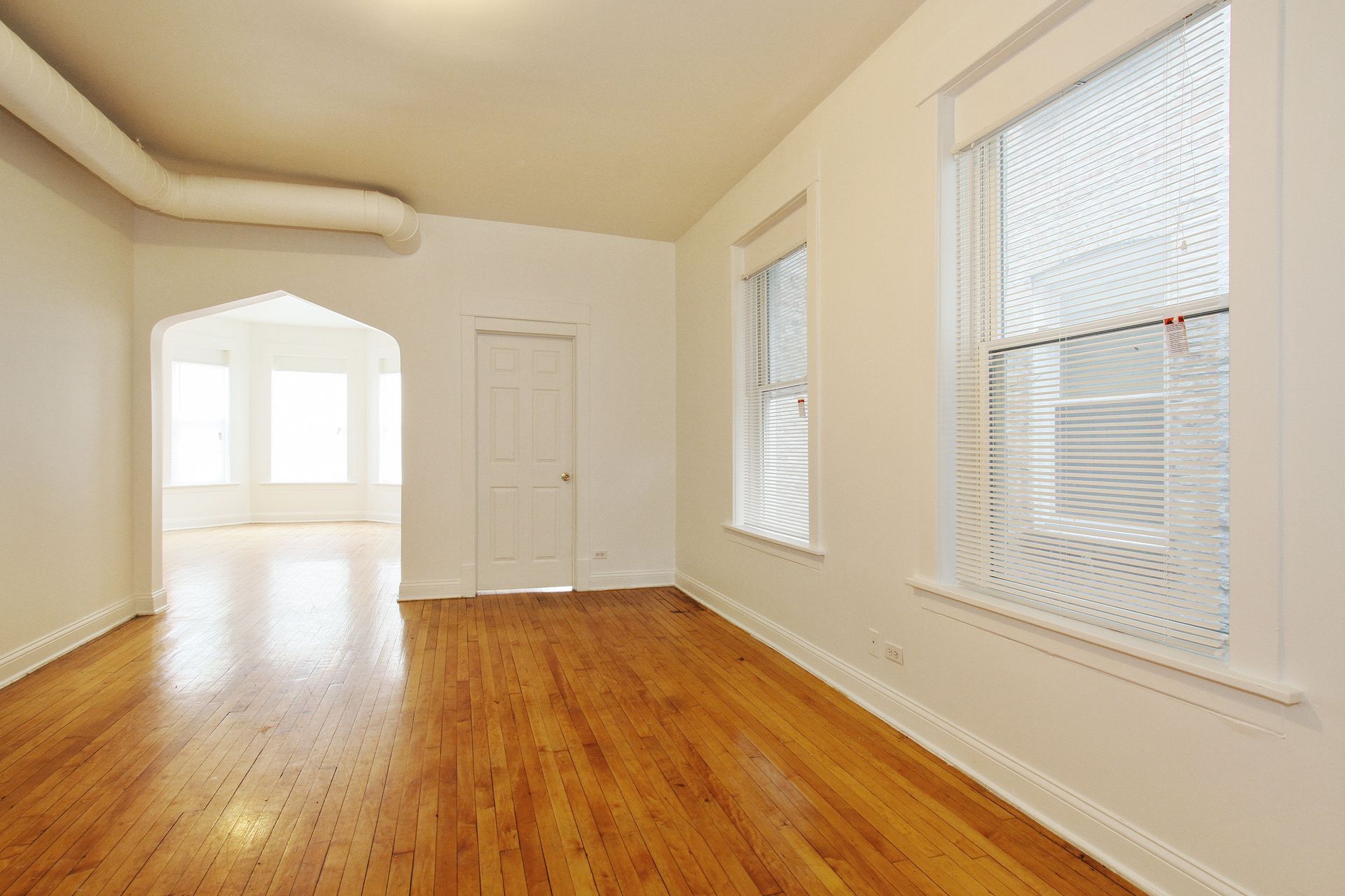 Empty living room with wood floors, two windows, and an arched doorway leading to another room.