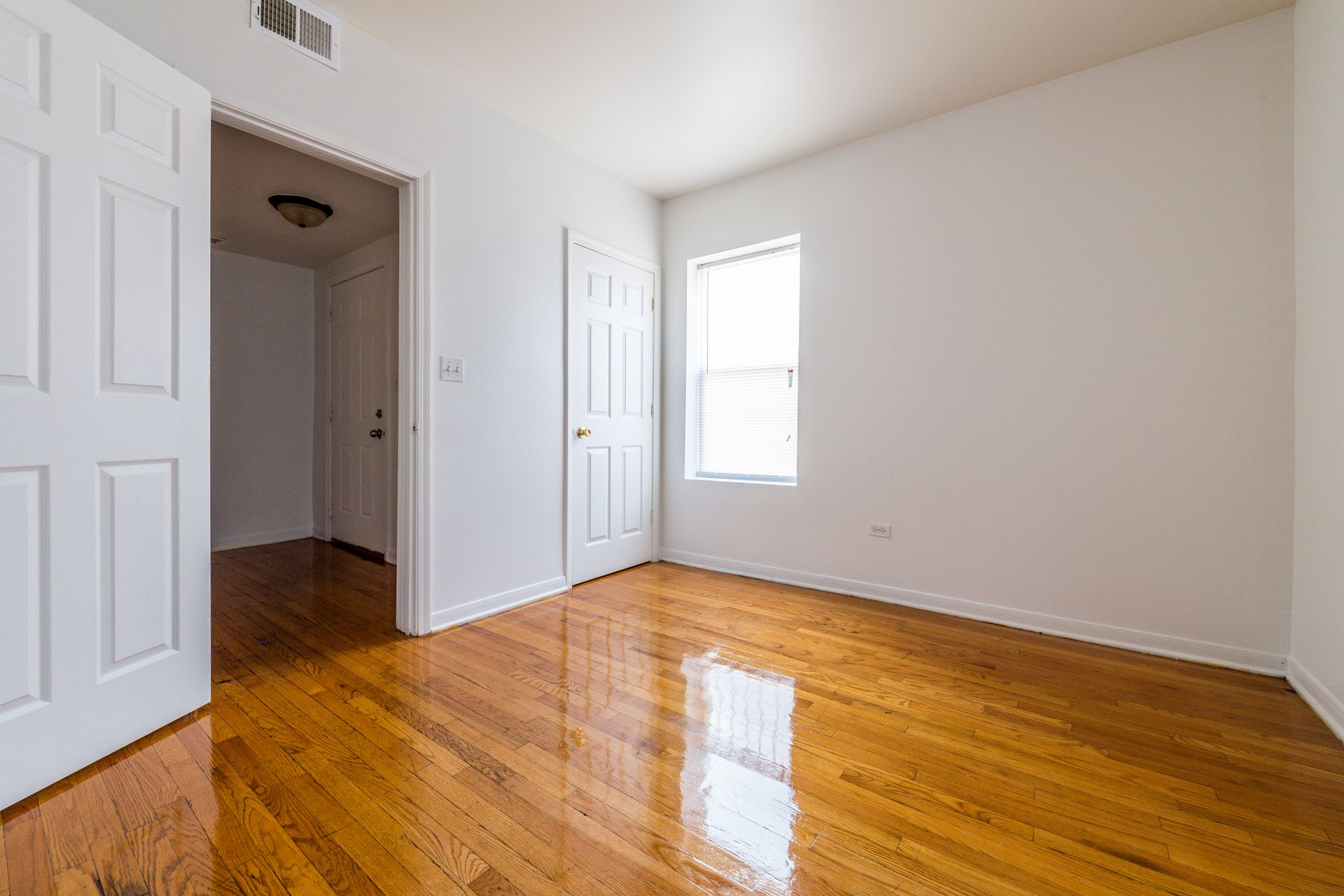 Empty room with hardwood floors, white walls, two closed doors, and a window.