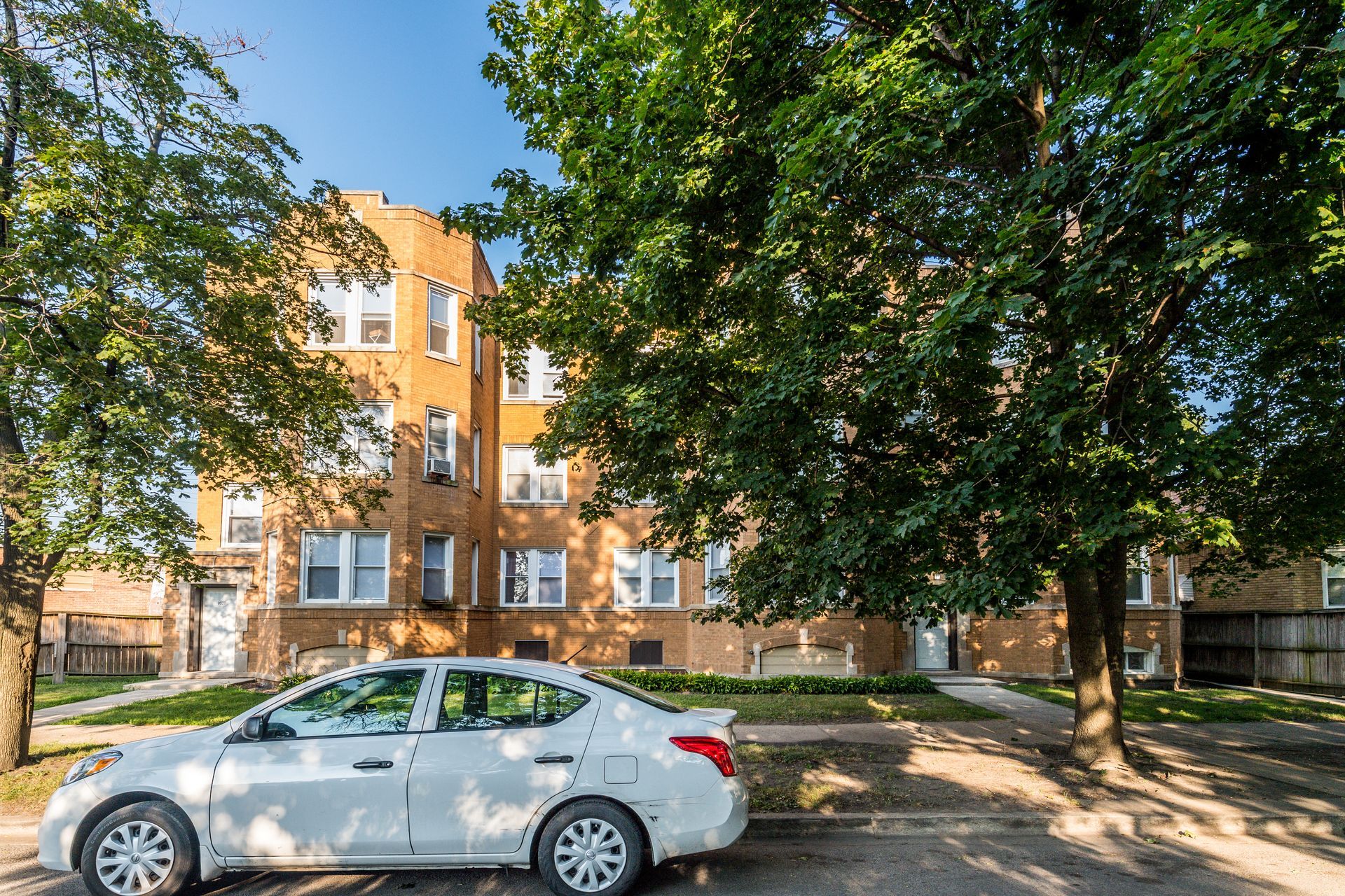 White car parked in front of a brick apartment building with trees and blue sky.