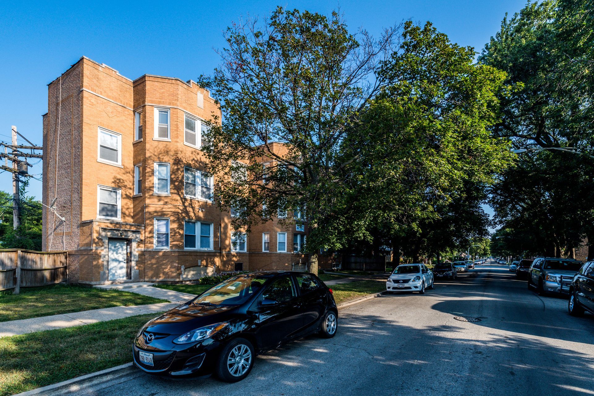 Brick apartment building on a tree-lined street with parked cars and a black car in the foreground.