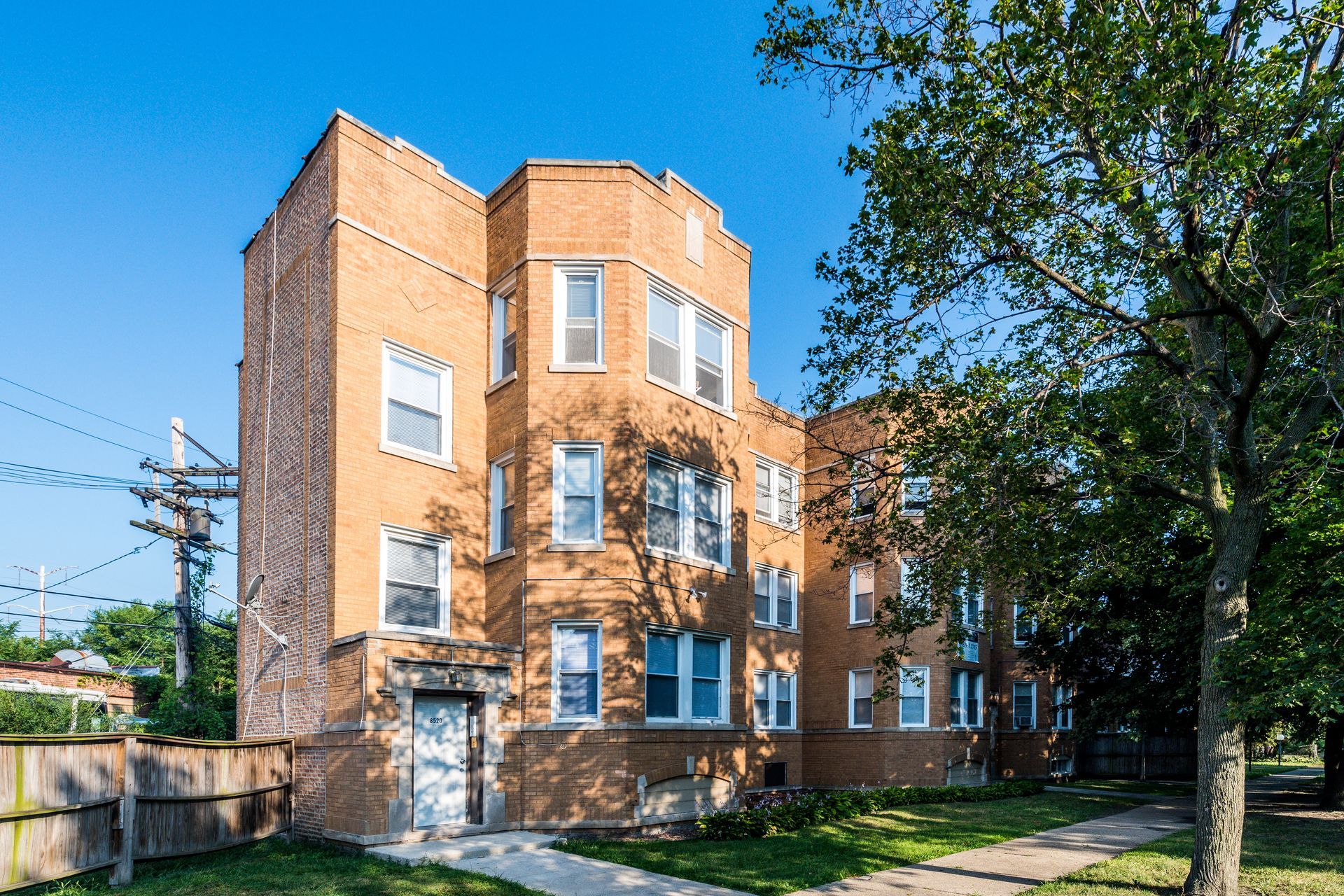 Three-story brick building with multiple windows, next to a tree and a wooden fence. Sunny day.