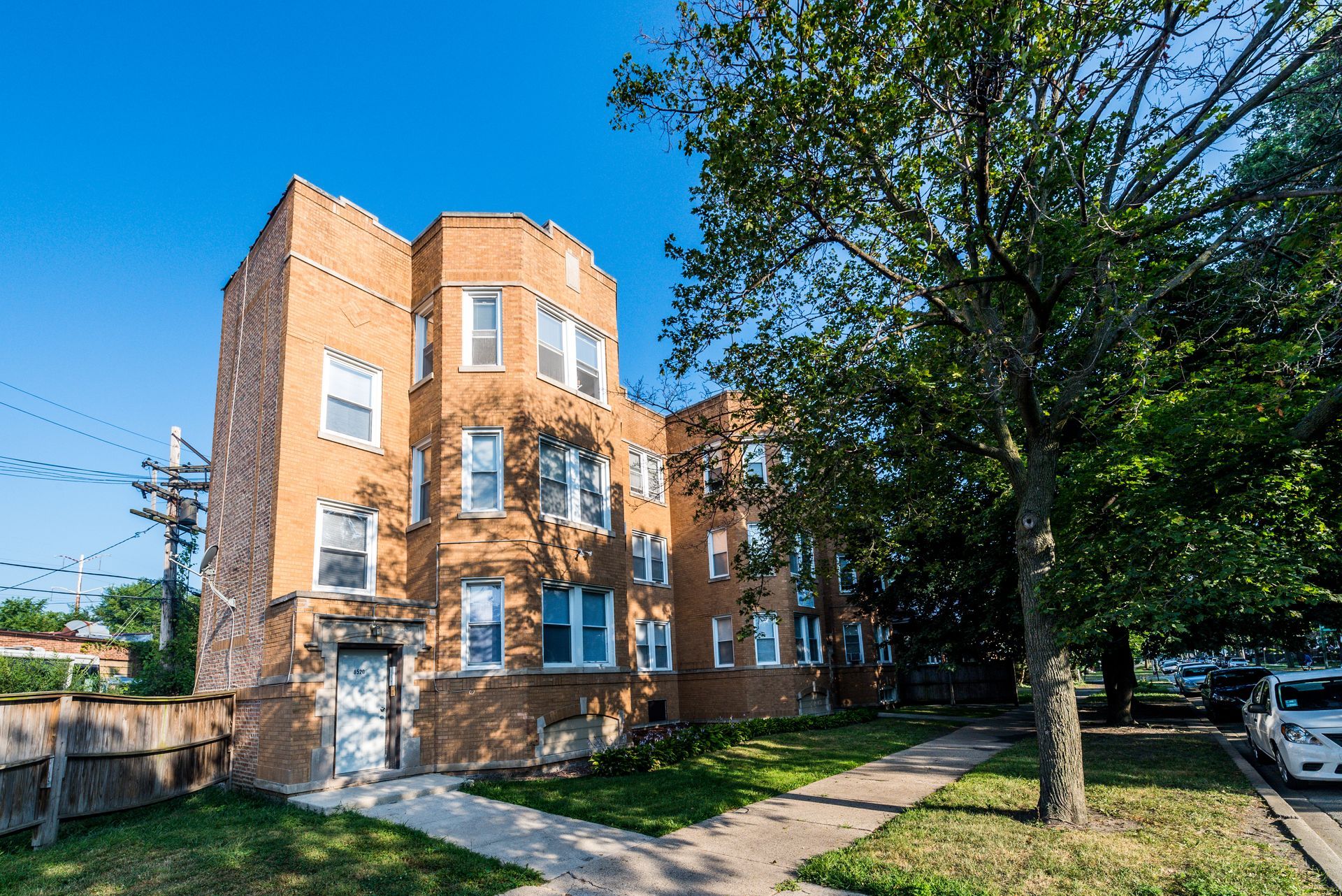 Multi-story brick apartment building with white-framed windows, a grassy yard, and a tree on a sunny day.