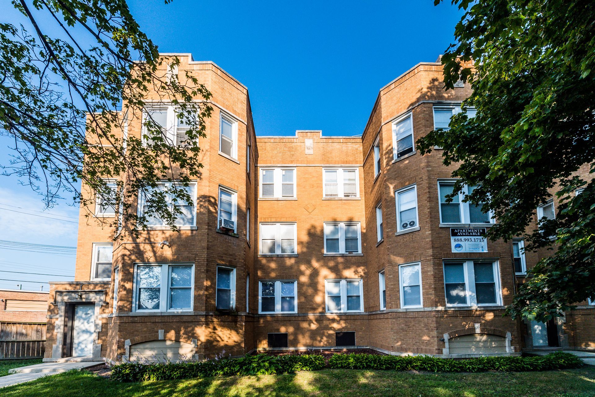 Brick apartment building with multiple windows under a blue sky, framed by trees and a patch of grass.