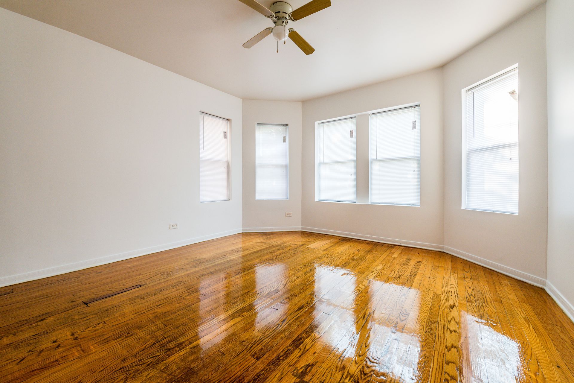 Empty room with hardwood floors, white walls, and a ceiling fan, with windows along a curved wall.