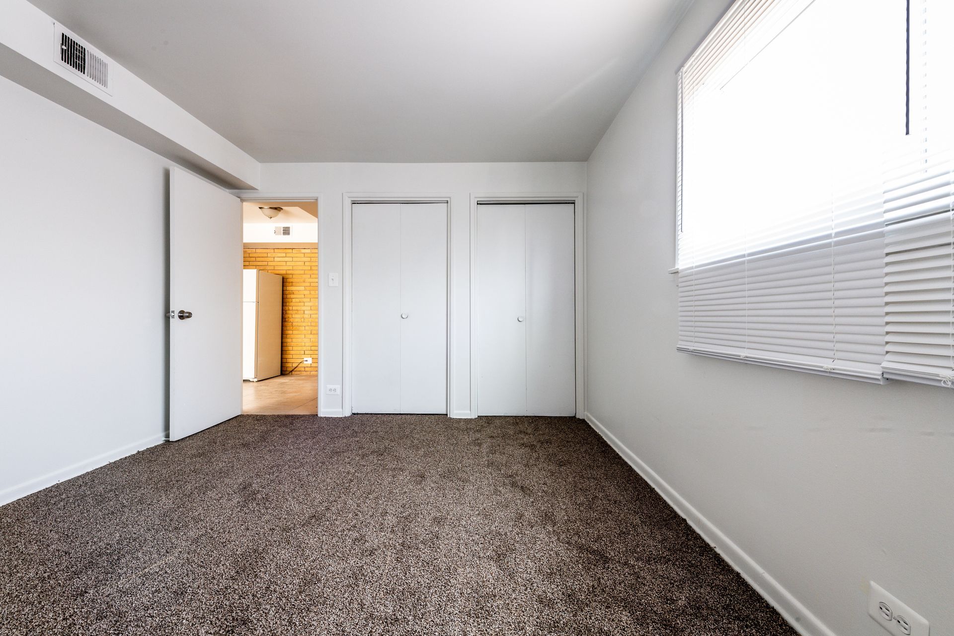 Empty bedroom with brown carpet, white walls, two closets, and a window with blinds.