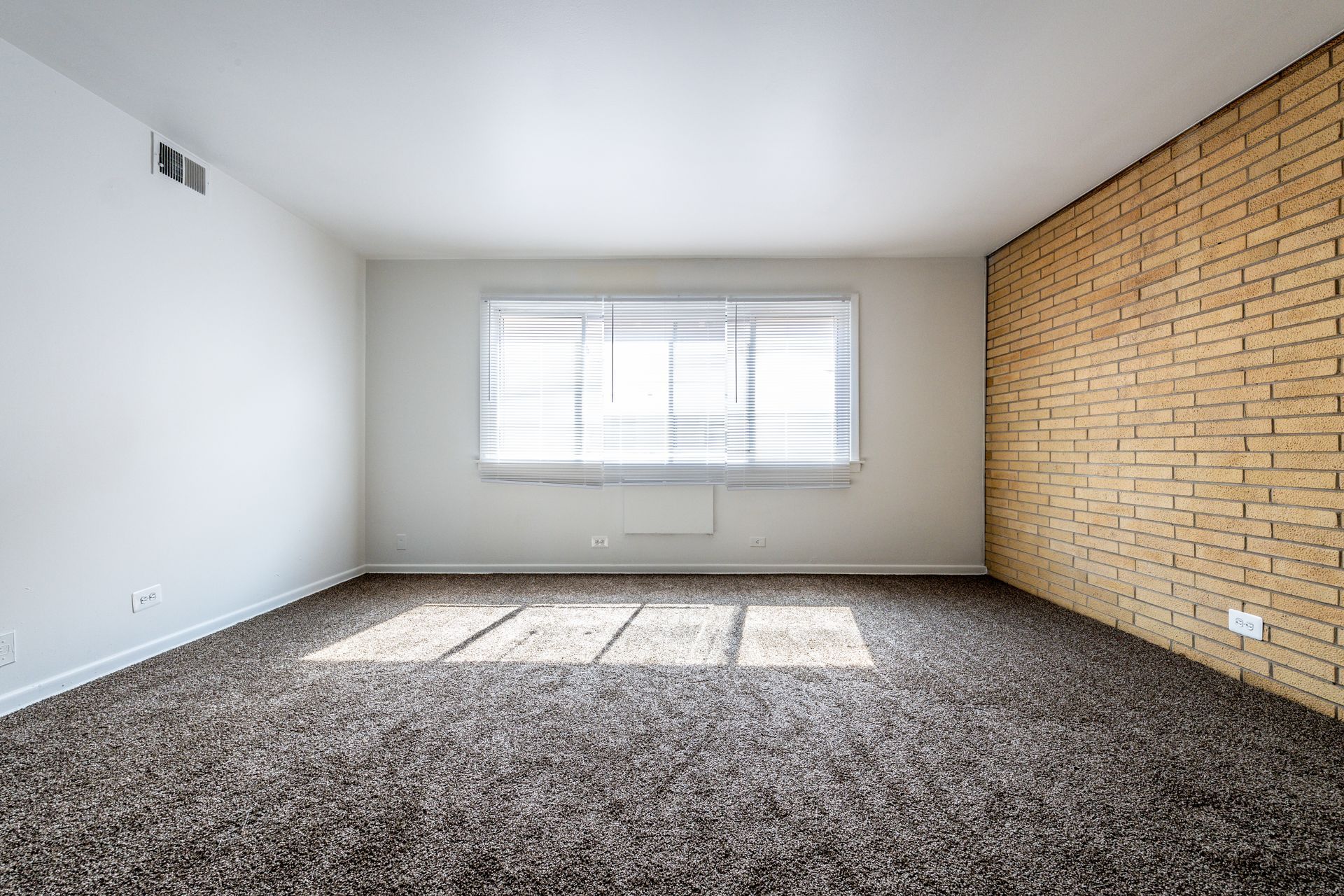 Empty room with brown carpet, window with light filtering, and a brick wall.