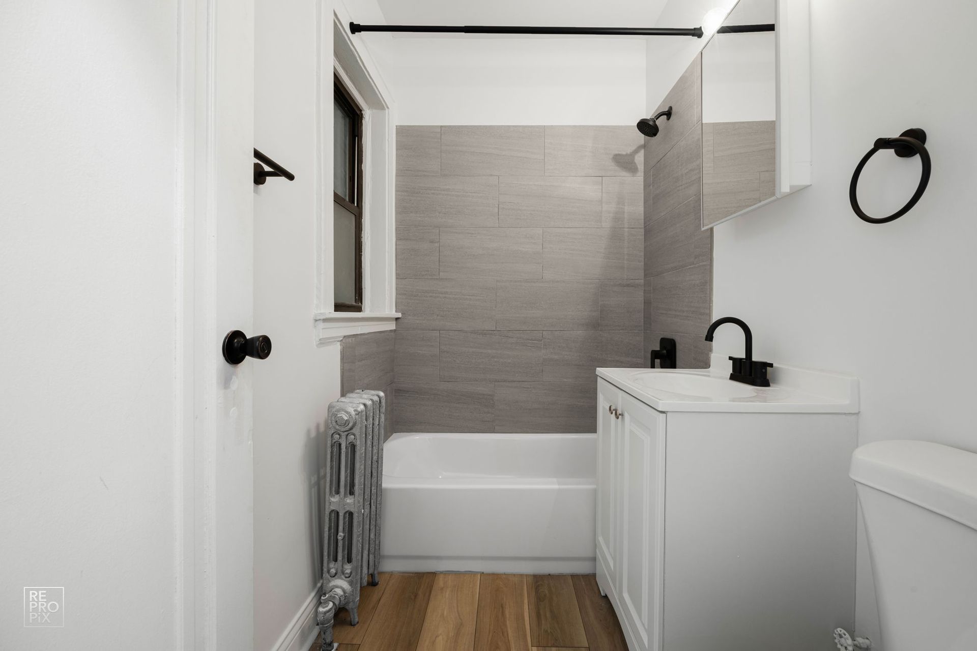 Small, white bathroom with gray tile. Features a white vanity, tub, toilet, and black fixtures.