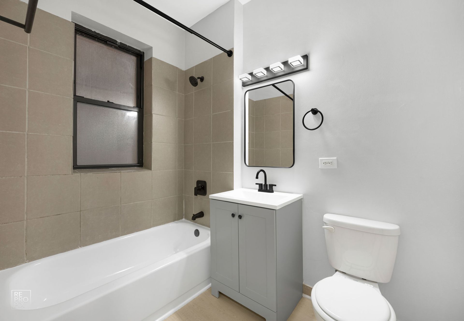 Bathroom with beige tile, white tub, gray vanity, black fixtures, and a window.