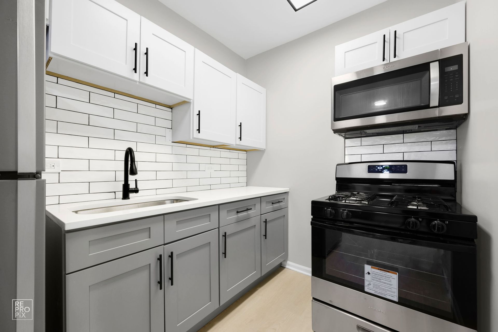 Modern kitchen with gray and white cabinets, stainless steel appliances, and white brick backsplash.