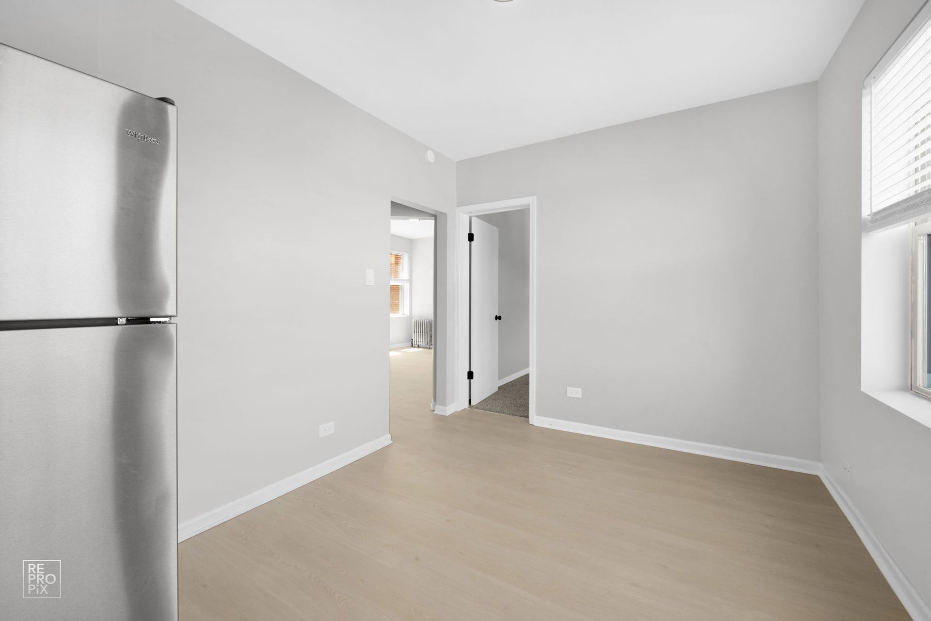 Empty room with light wood floor, gray walls, stainless steel fridge, and doors.