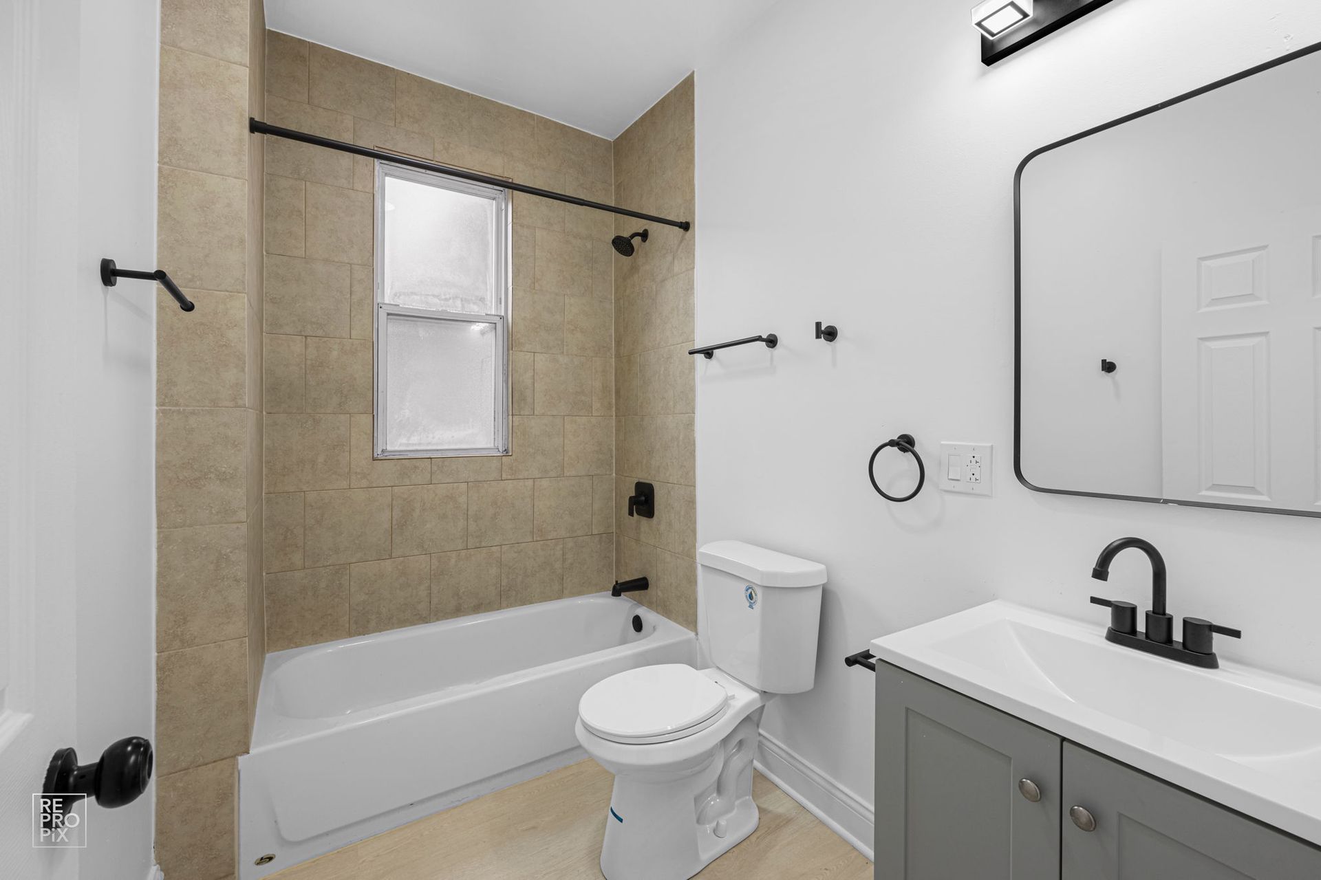 Bathroom with a white tub and toilet, gray vanity, tan tile, and black fixtures.