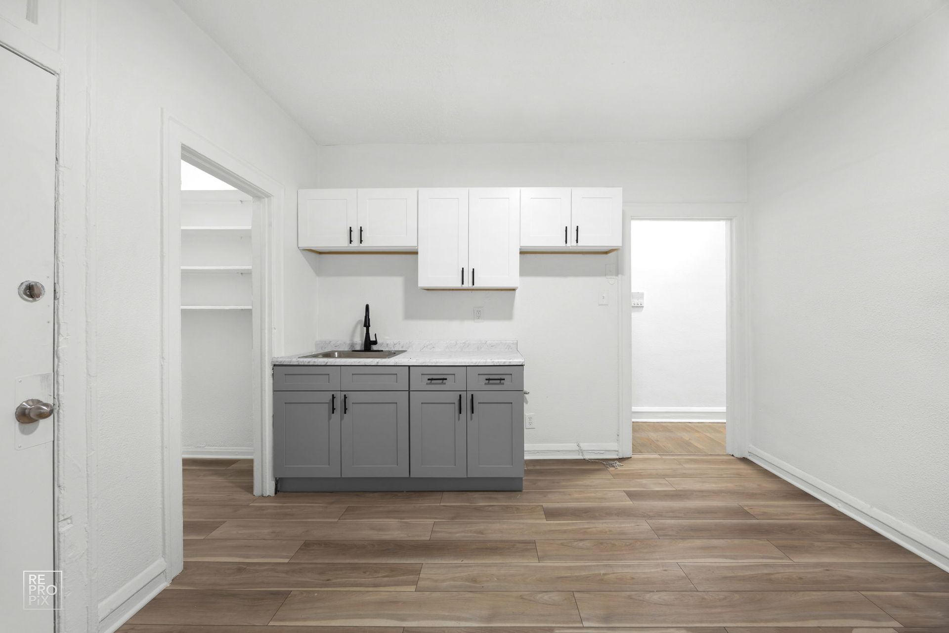 Small kitchenette with gray and white cabinets, sink, and wooden floor. Open doorways lead to storage and another room.
