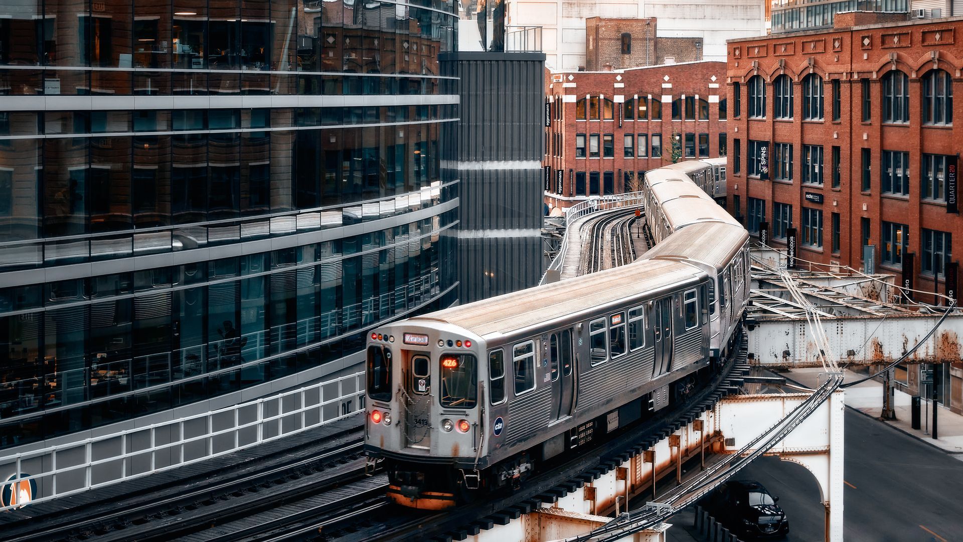 Elevated train curving past modern glass building and old brick structures in city.