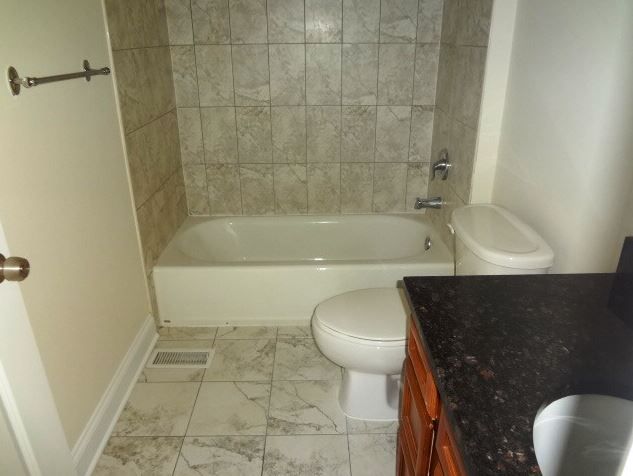 Bathroom with a white bathtub, toilet, and marble-like tile flooring and wall. A brown vanity is in the foreground.