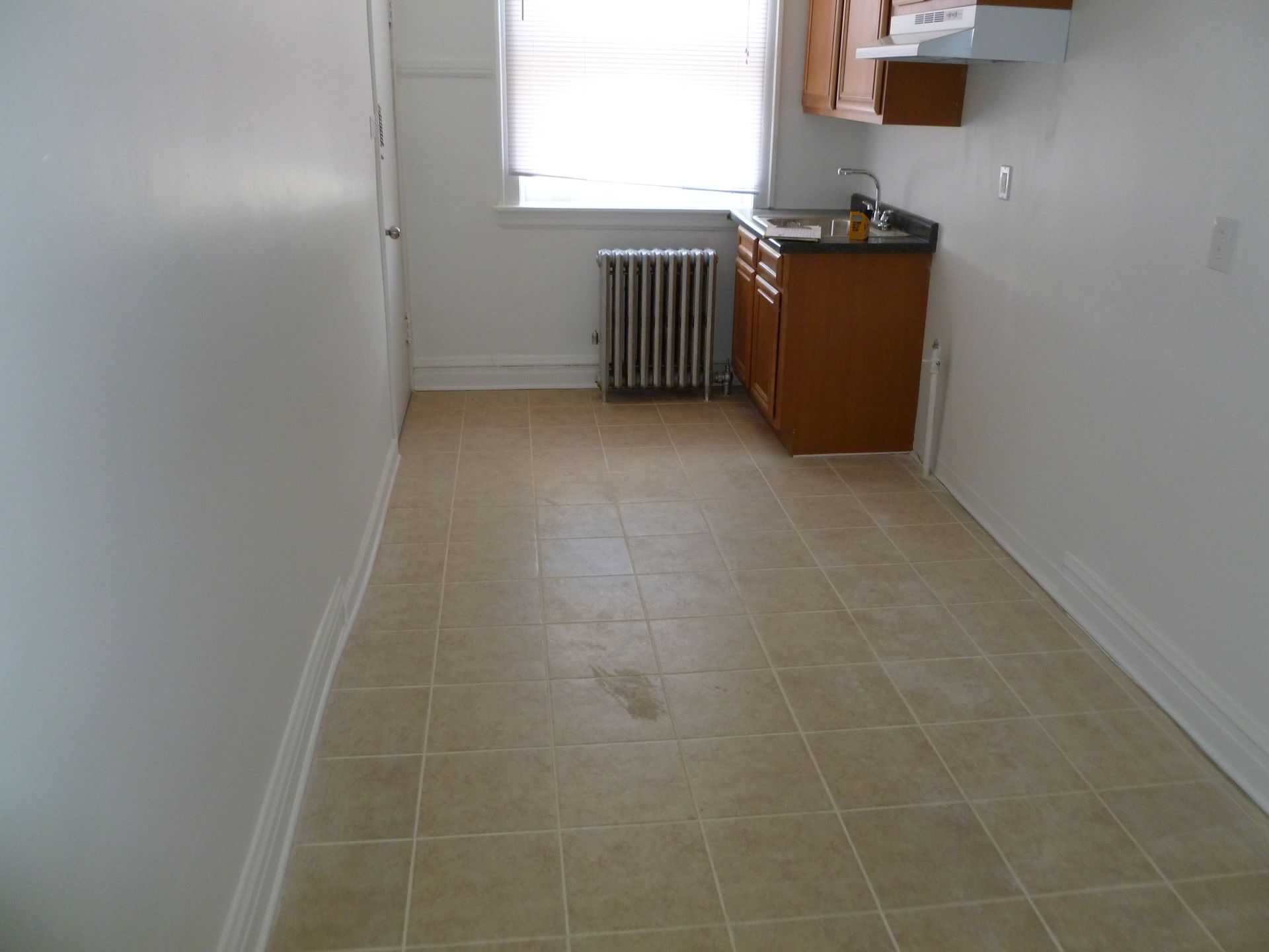 Small, empty kitchen with tan tile floor, wooden cabinets, and a radiator.