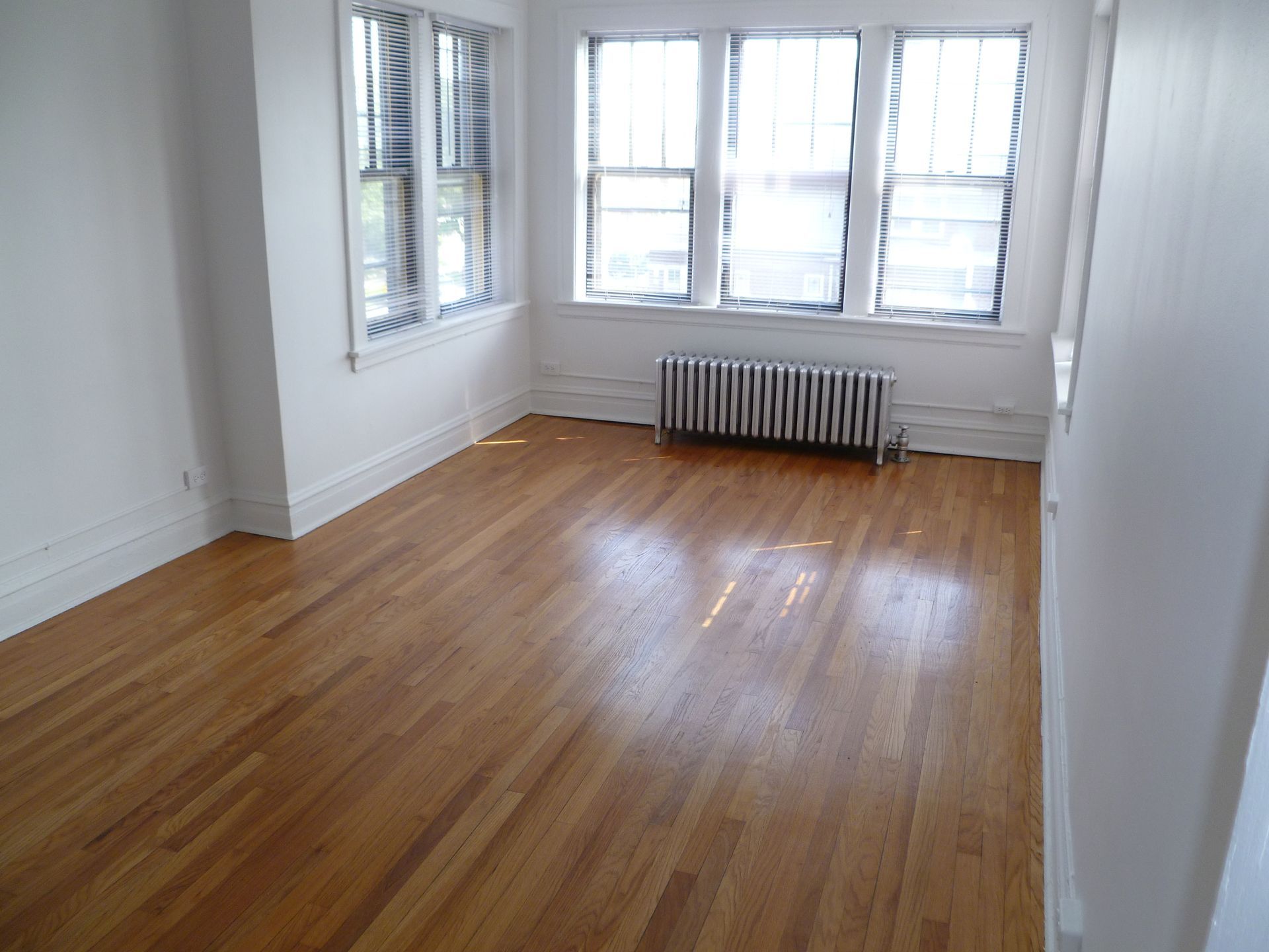 Empty room with hardwood floors, white walls, and three windows with radiator beneath.