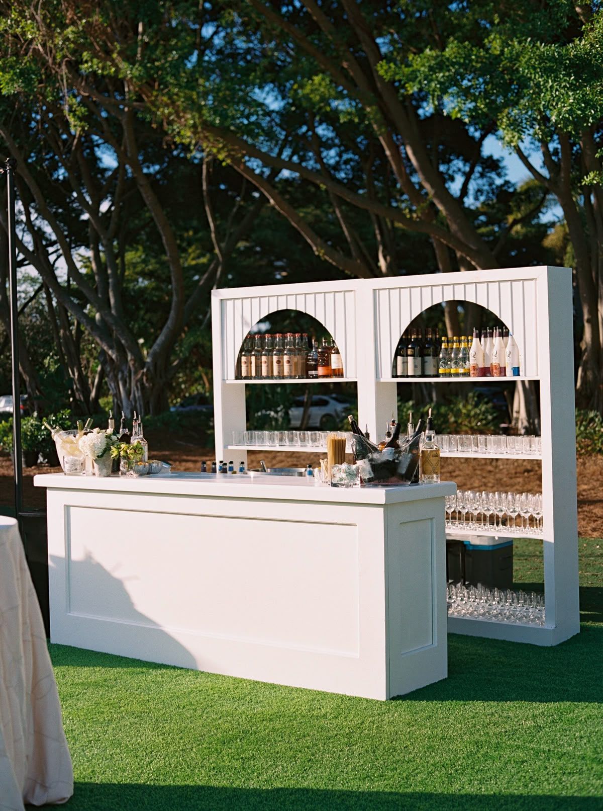 A white, wooden portable bar with arched shelving displaying liquor bottles and glassware, set up on a grassy lawn.
