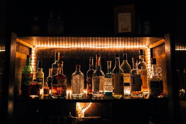 A dimly lit bar shelf illuminated by warm LED strip lights, displaying a collection of various liquor bottles.