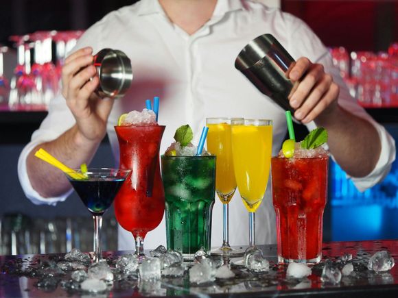 A bartender in a white shirt shakes a cocktail tin behind a counter featuring five colorful drinks with ice and straws.