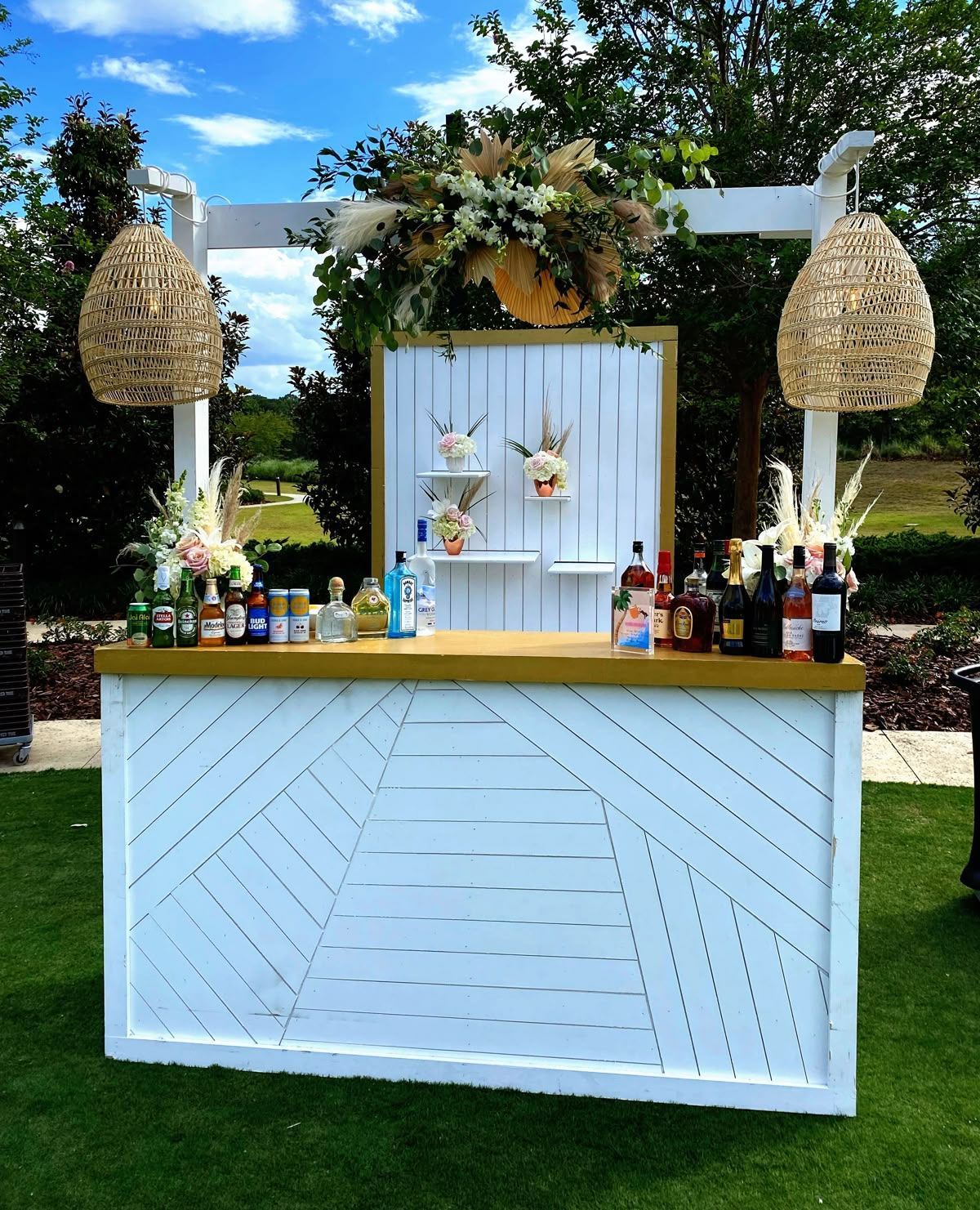A white wooden outdoor bar with a gold top, featuring floral arrangements and hanging wicker pendant lights on a lawn.