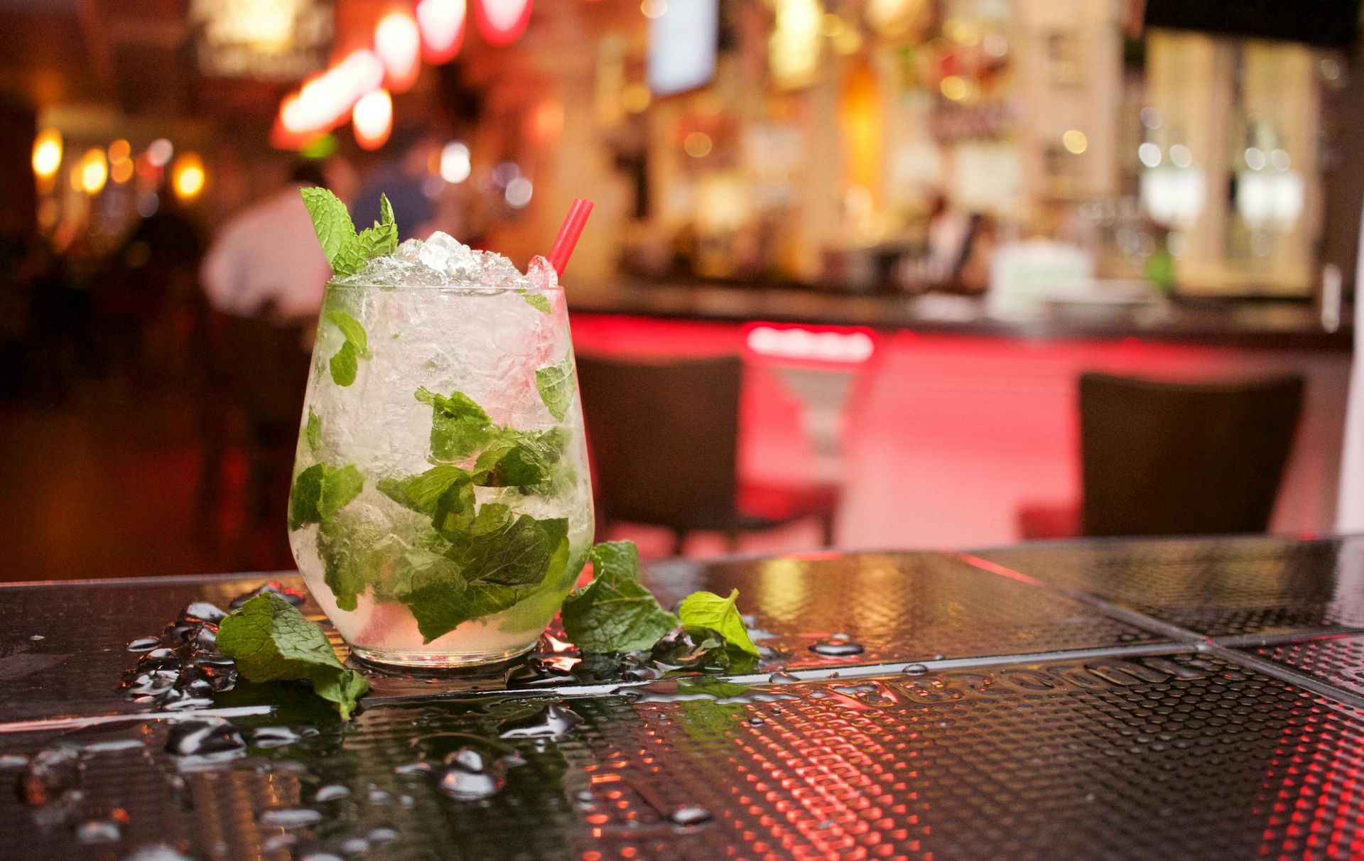 A cold mojito cocktail garnished with fresh mint on a reflective bar counter, with a blurred bar setting in the background.