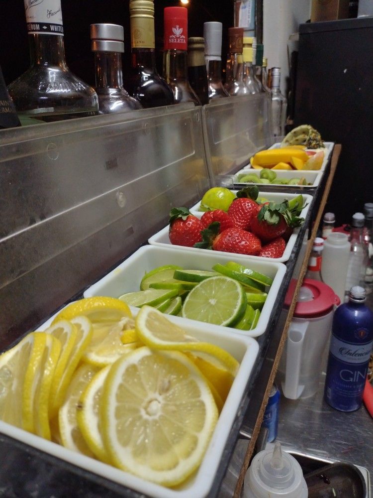 A bar garnish station with white containers holding sliced lemons, limes, strawberries, and other fruits.