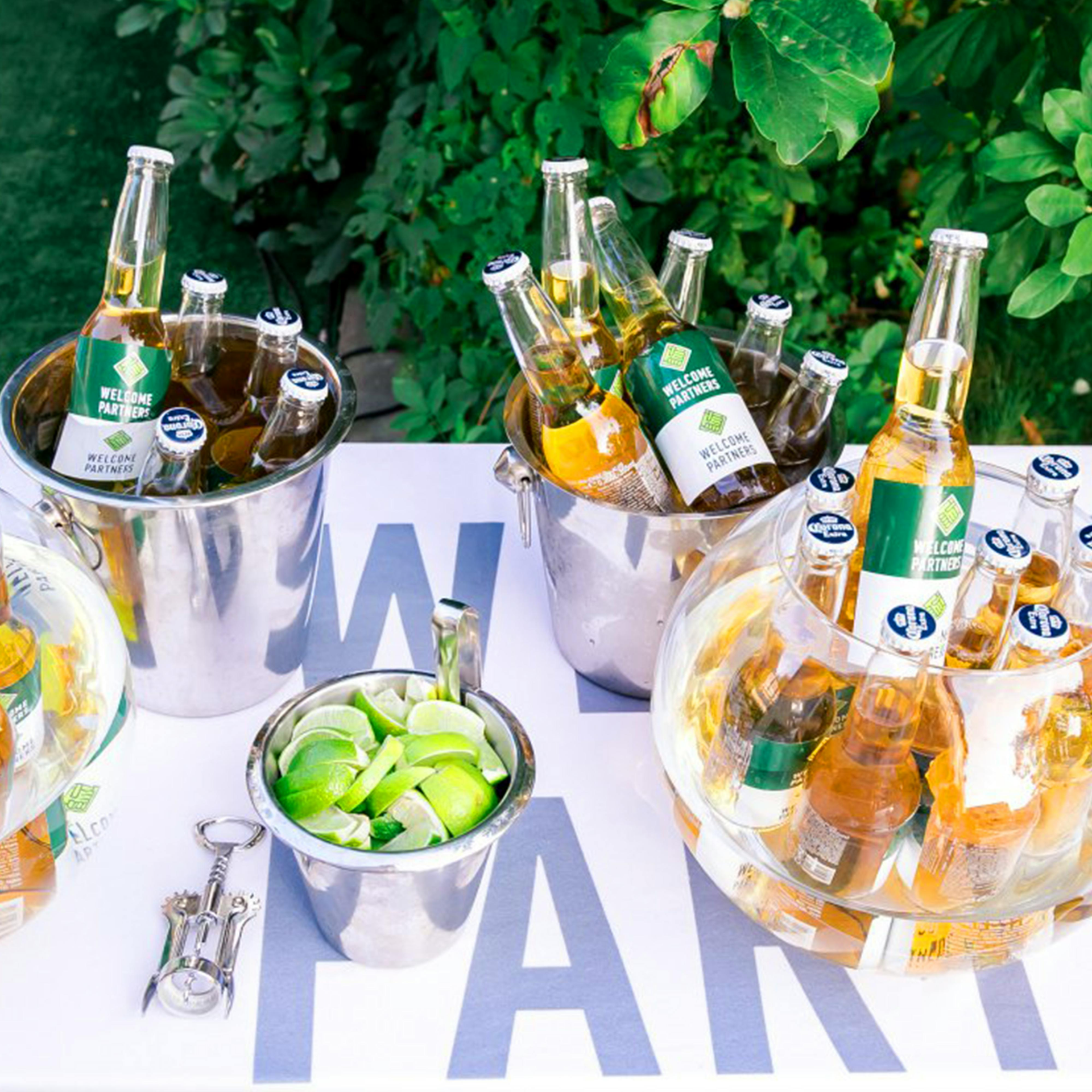 Buckets of chilled beer and a small bowl of lime wedges are arranged on a party table with a bottle opener.