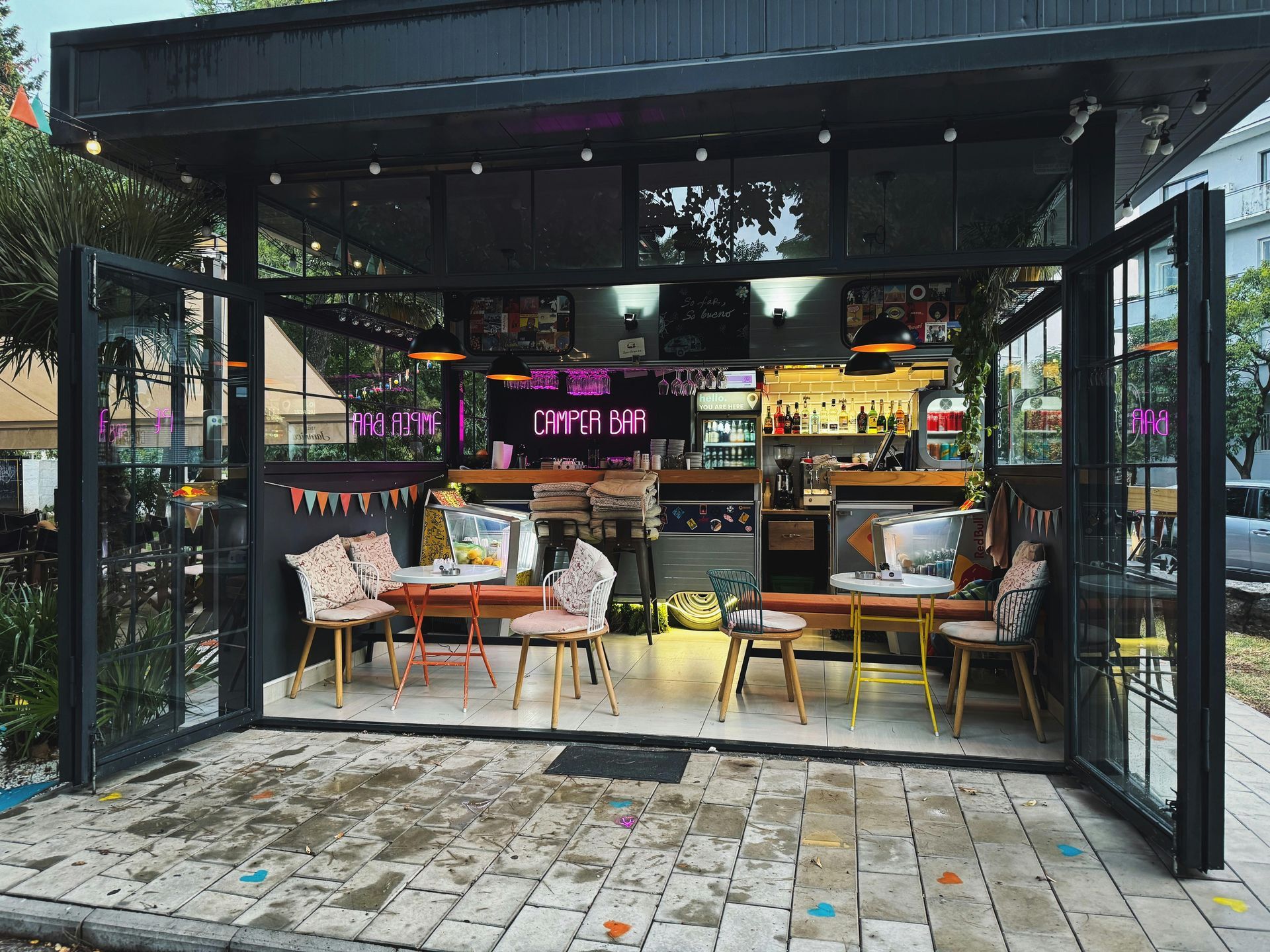 A small, open-fronted cafe with metal framing, interior tables, chairs, and warm lighting on a paved patio.