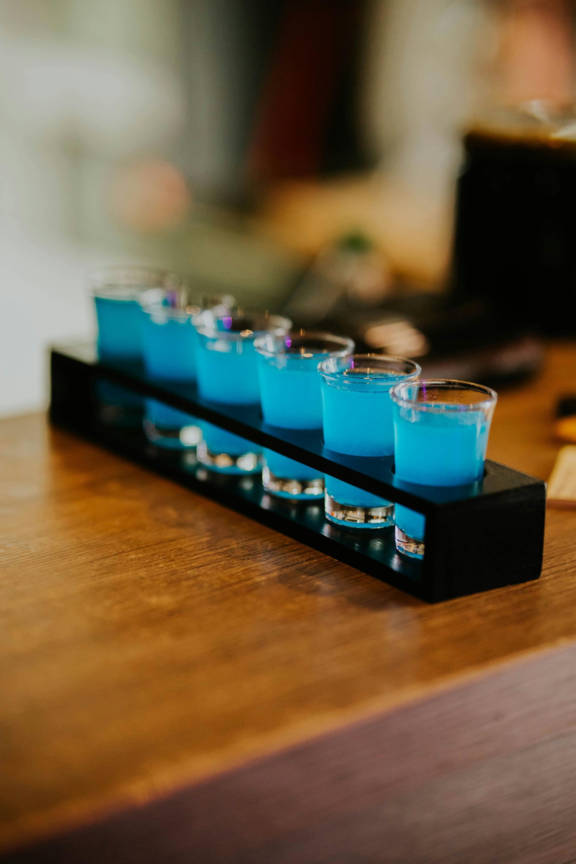 A row of six shot glasses filled with bright blue liquid sits in a black wooden holder on a wooden bar top.