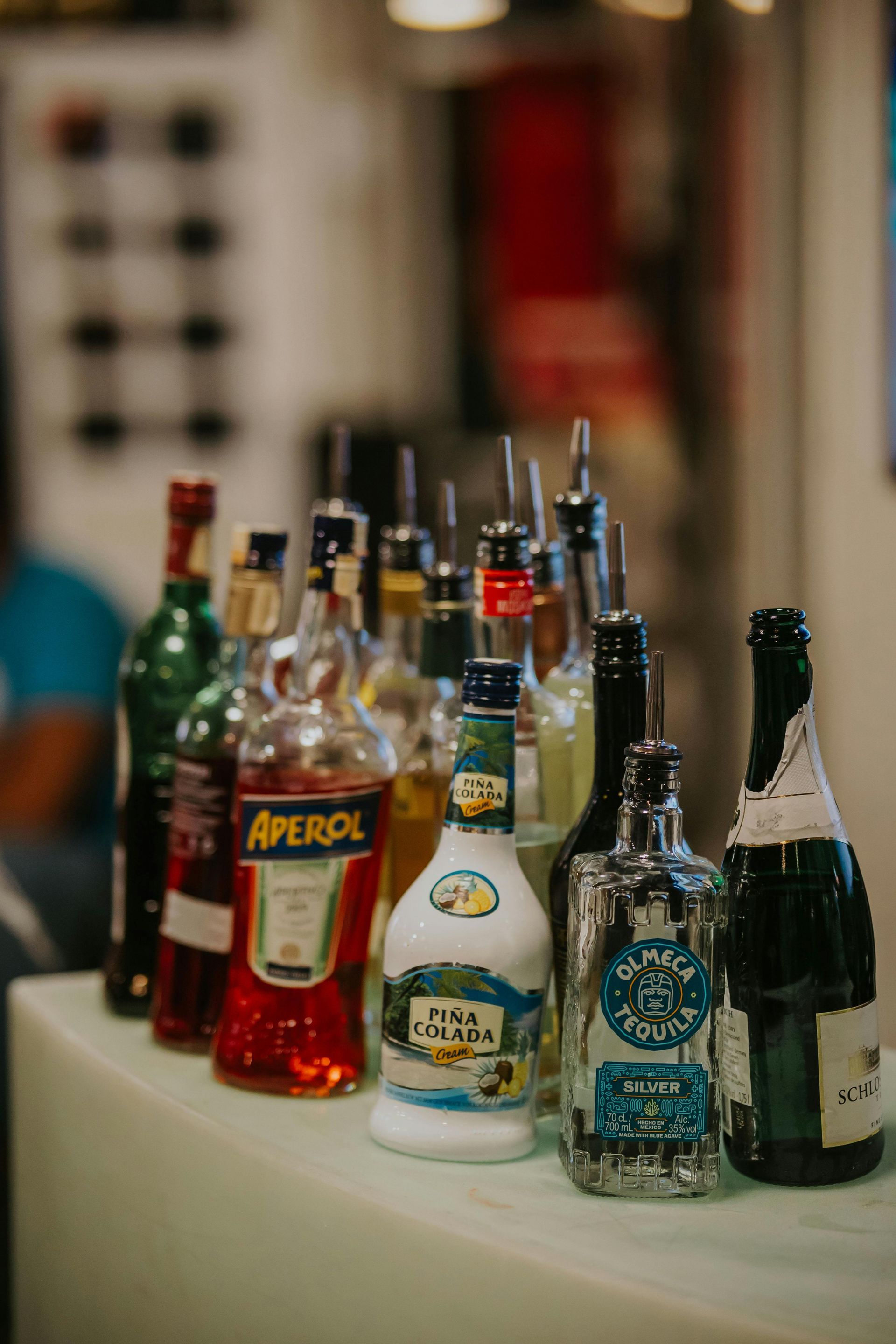 A line of assorted liquor bottles, including Aperol and Olmeca tequila, arranged on a bar counter.